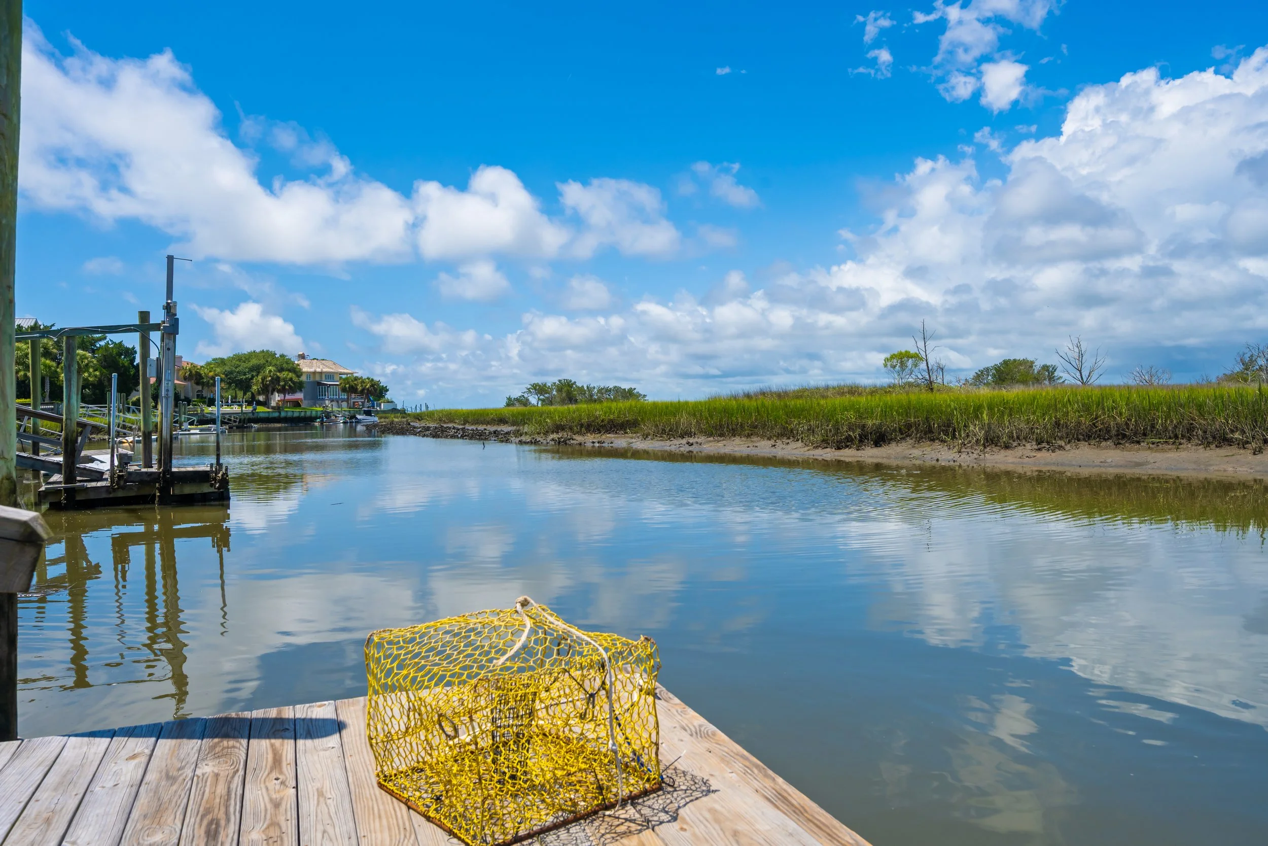 A dock on a river with a yellow fishing net in the foreground, calm water reflecting the blue sky and white clouds, houses with boats on the bank, and green grass and trees along the riverbank.