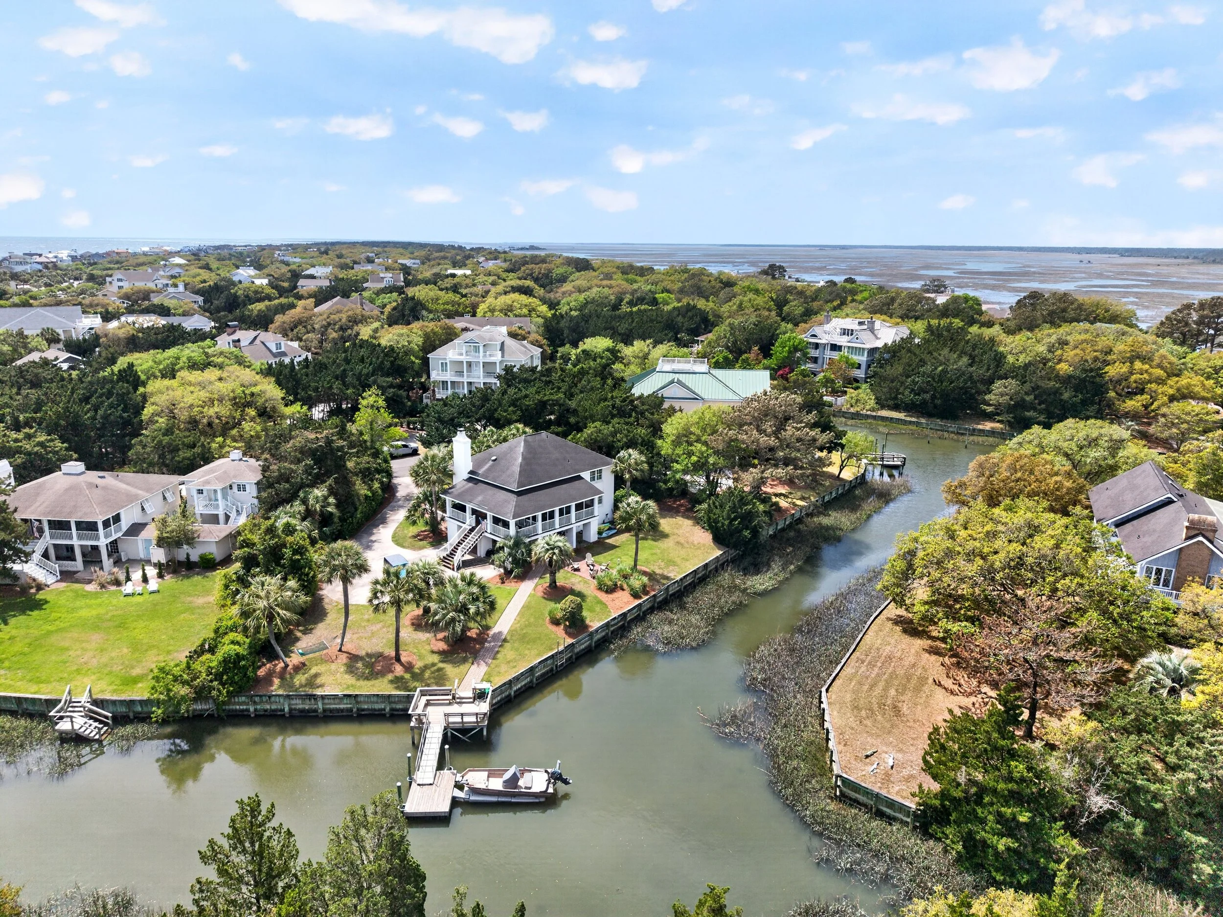 Aerial view of a coastal residential area with houses surrounded by lush green trees, a winding canal with a dock and boat, and marshlands in the background under a partly cloudy sky.