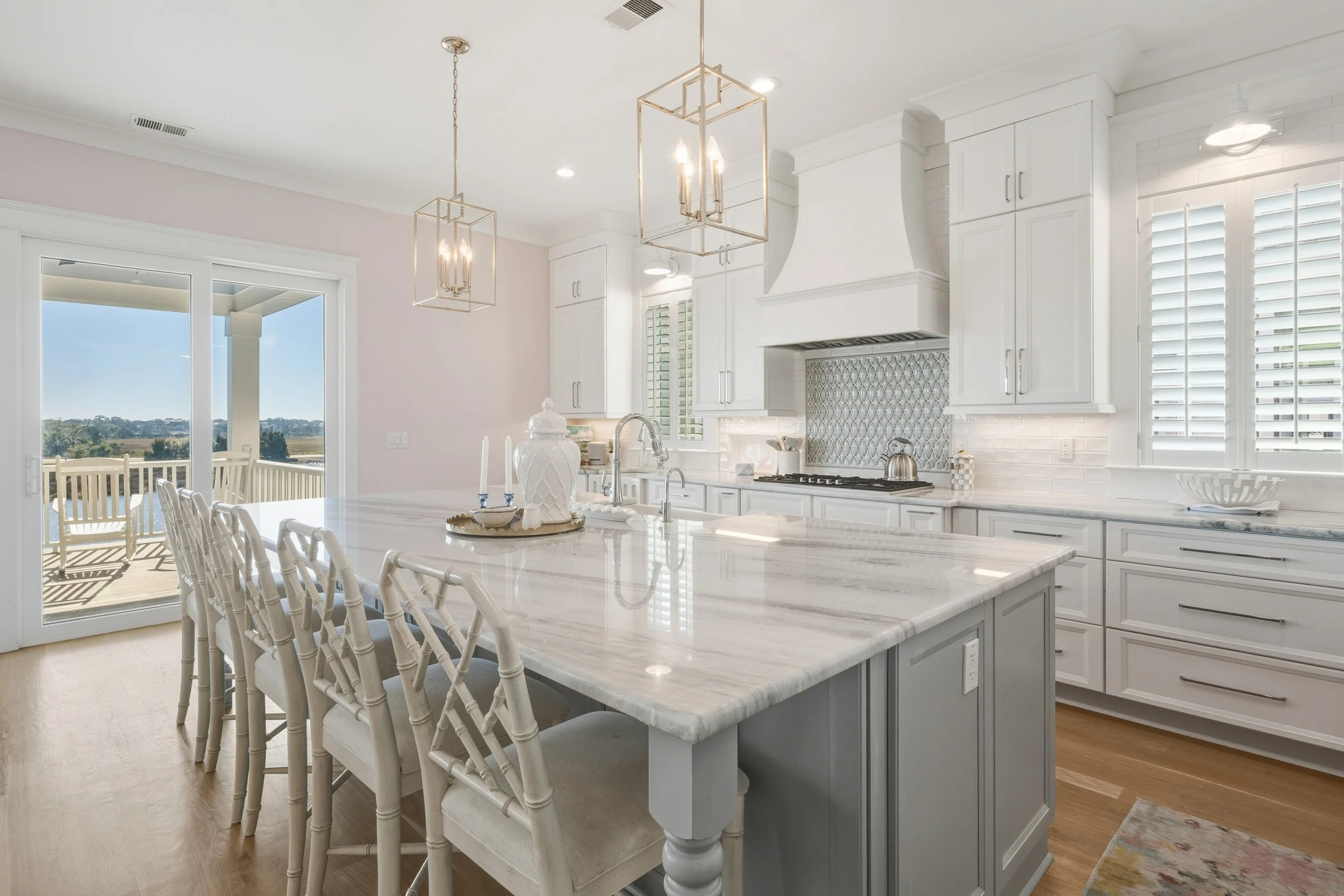 Bright, modern kitchen with white cabinetry, marble island, and pendant lights, opening to a balcony with outdoor seating and scenic view.