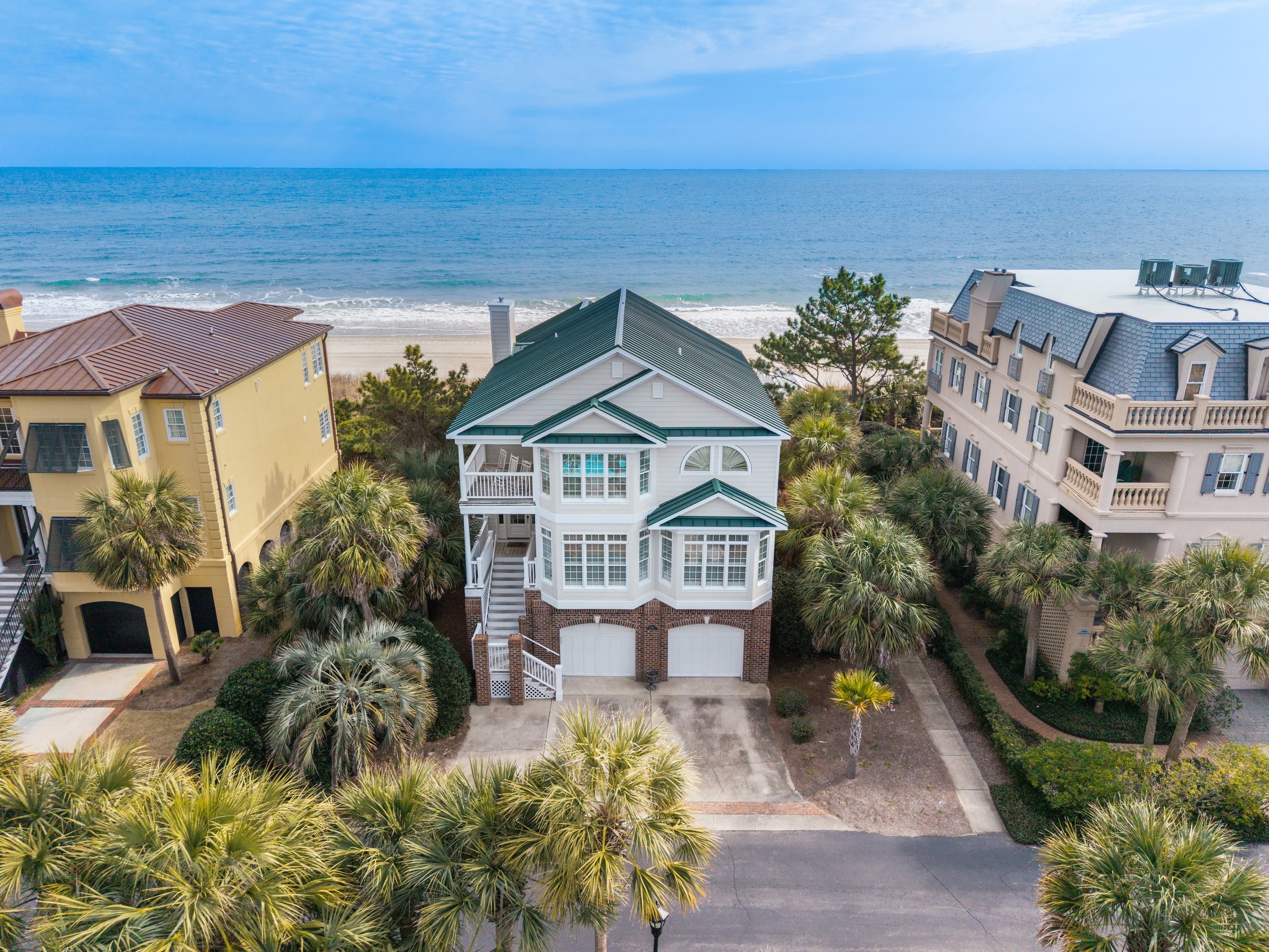 A beachfront scene with multi-story houses surrounded by palm trees, overlooking the ocean with a cloudy sky.