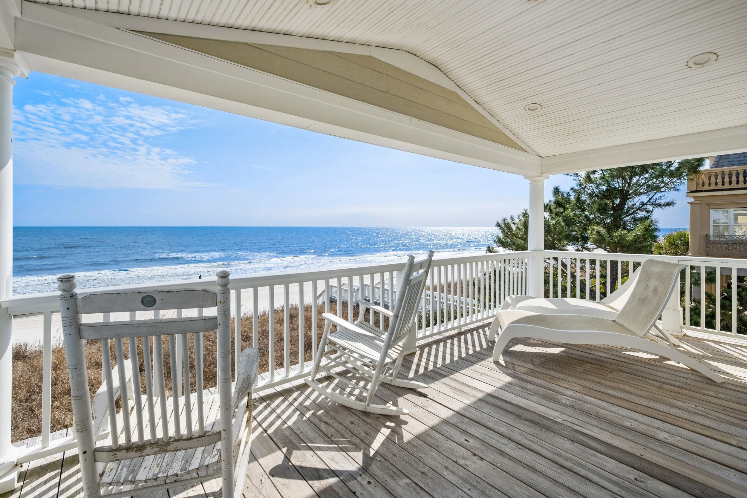 View from a porch overlooking the ocean with white chairs and railing, sunny sky, and a few trees.