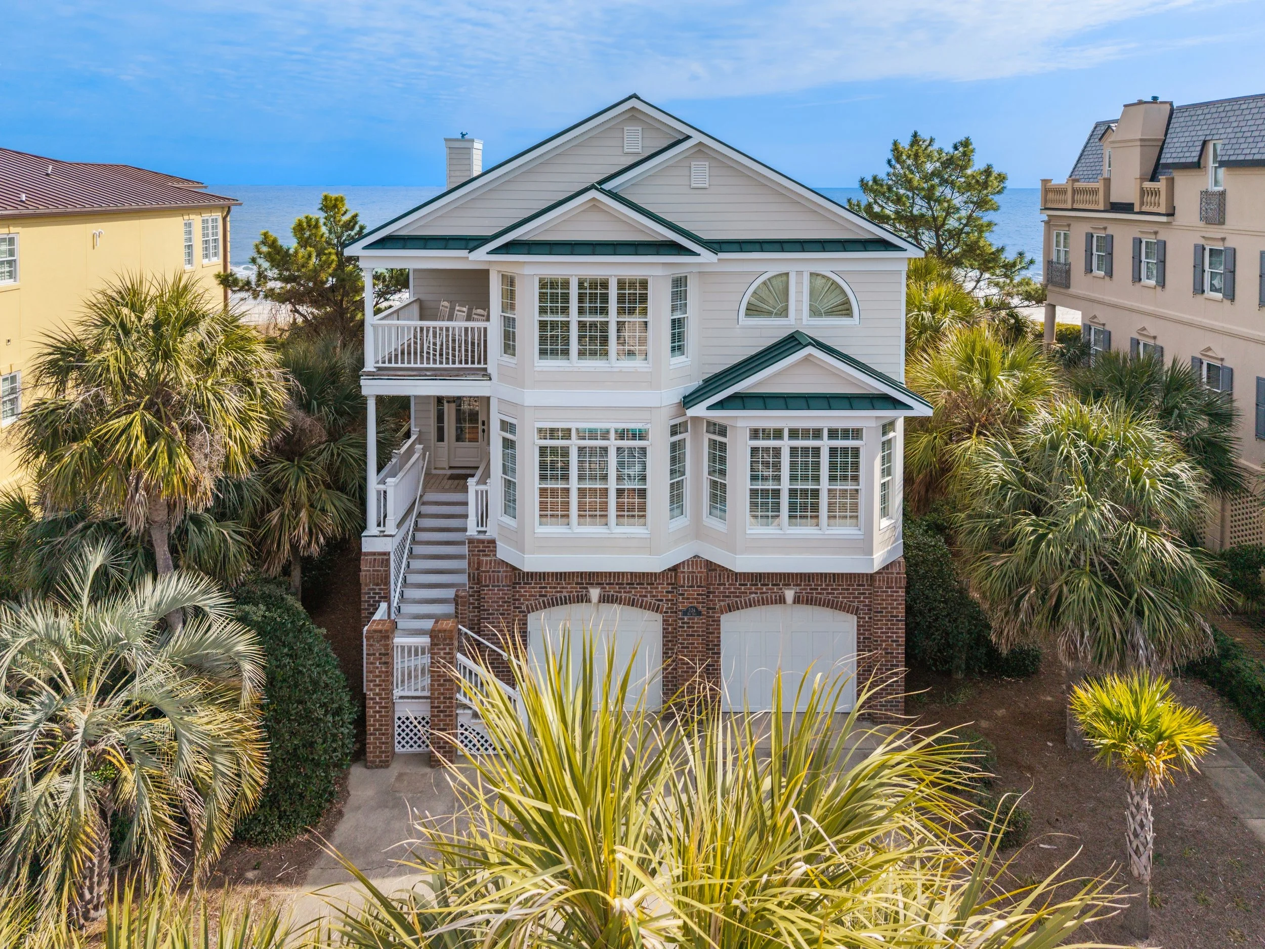 A white, multi-story house with green roofing and large windows, surrounded by palm trees and vegetation, with the ocean and sky visible in the background.