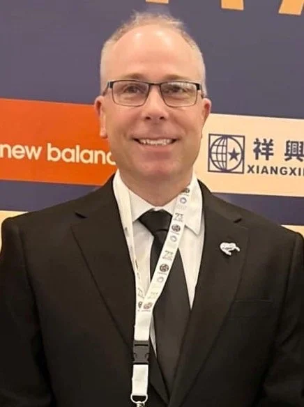 Andrew Collings in a black tuxedo with glasses and a lanyard, smiling at a professional or conference event.