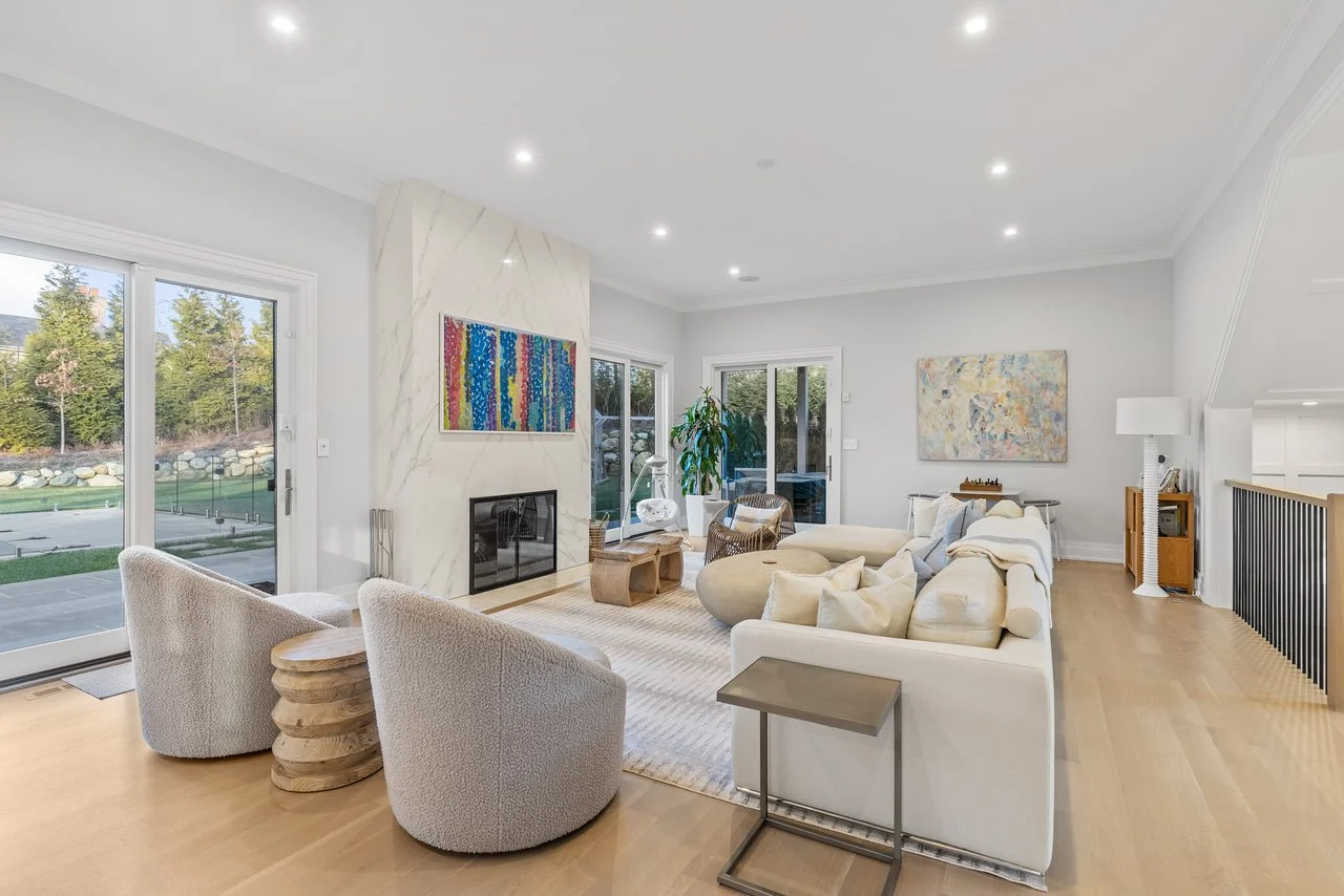 Modern living room with white walls, hardwood floors, white sectional sofa, armchairs, fireplace, large windows, abstract artwork, potted plants, and outdoor view.