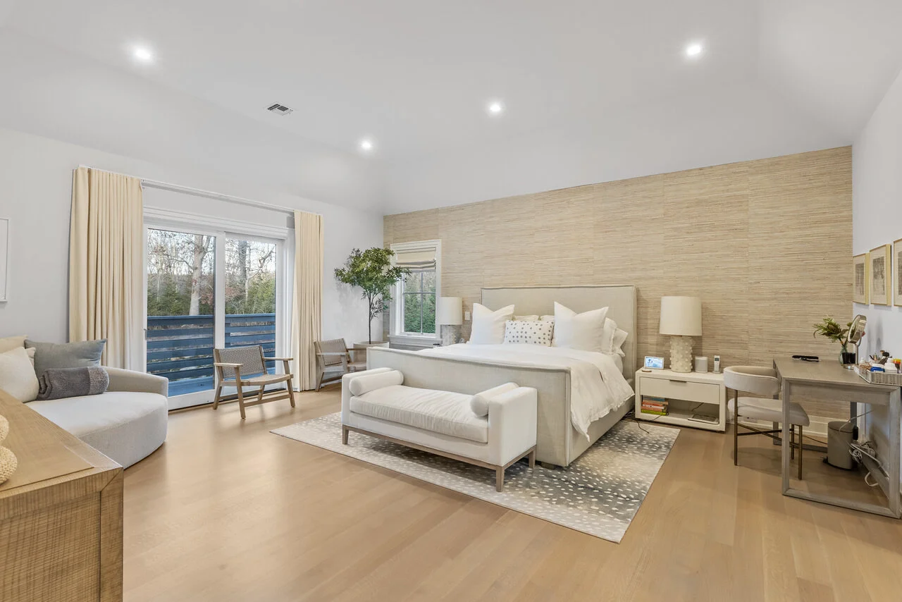 Bright, modern bedroom with a large bed, white and neutral tones, wooden accents, a seating area, and large windows with view of trees.