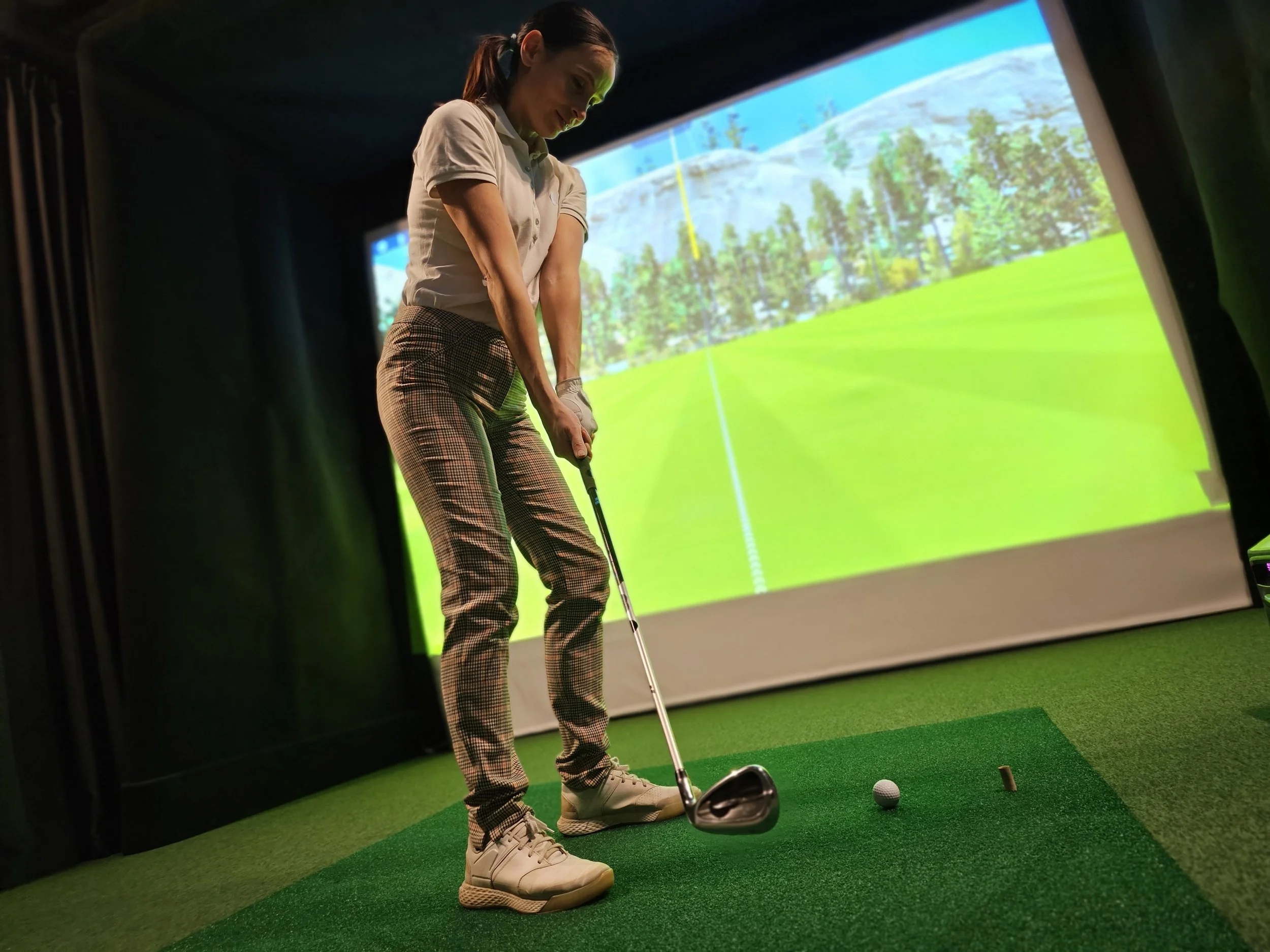 A woman preparing to hit a golf ball on an indoor golf simulation with a projected landscape of trees and mountains in the background.