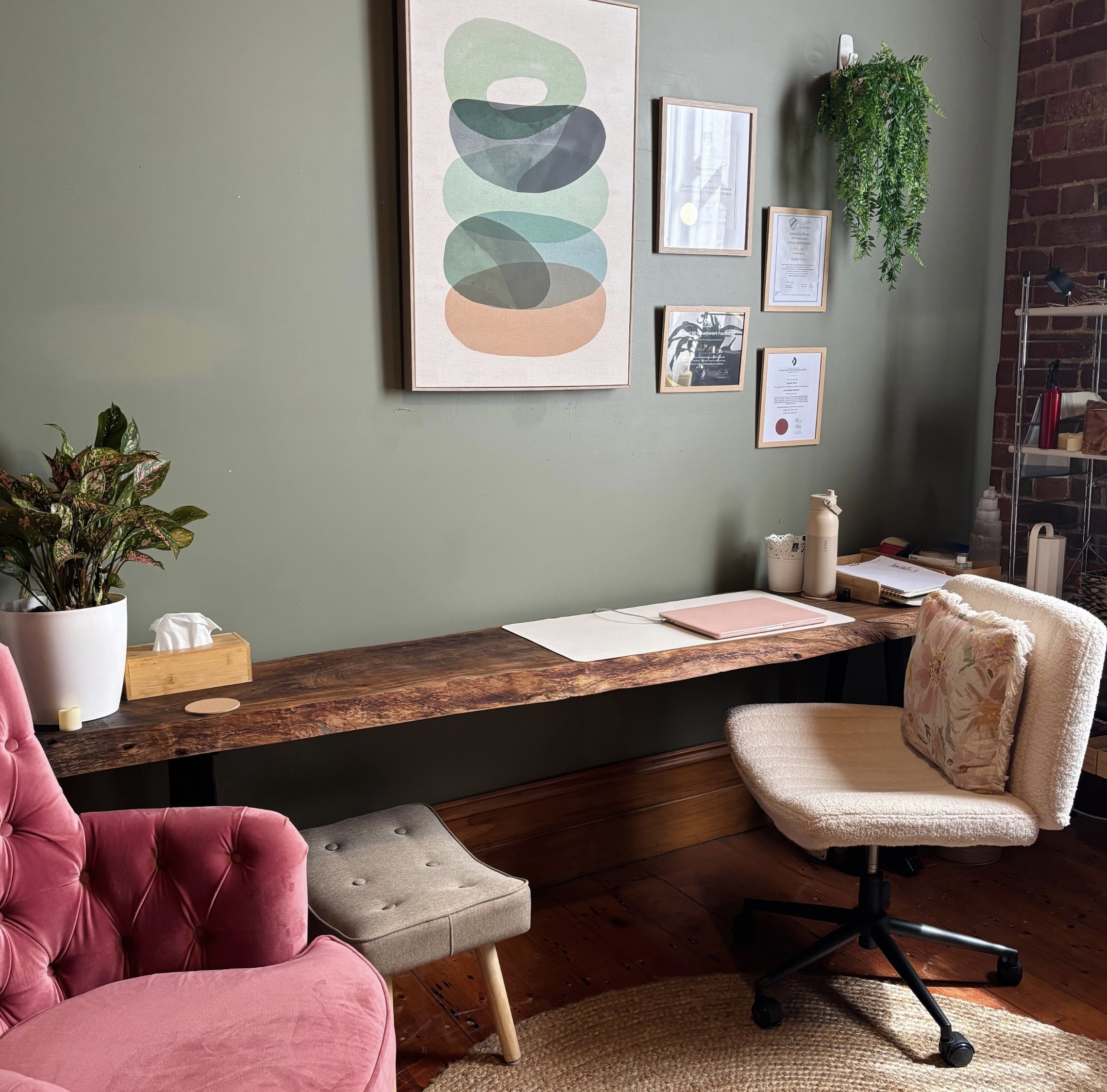 A cozy workspace with a wooden desk against a green wall decorated with framed artwork and certificates. There is a white office chair with a floral cushion, a pink velvet chair, and a small upholstered footstool. On the desk are a potted plant, tissues, water bottles, and office supplies. A hanging plant is on the right side of the wall.