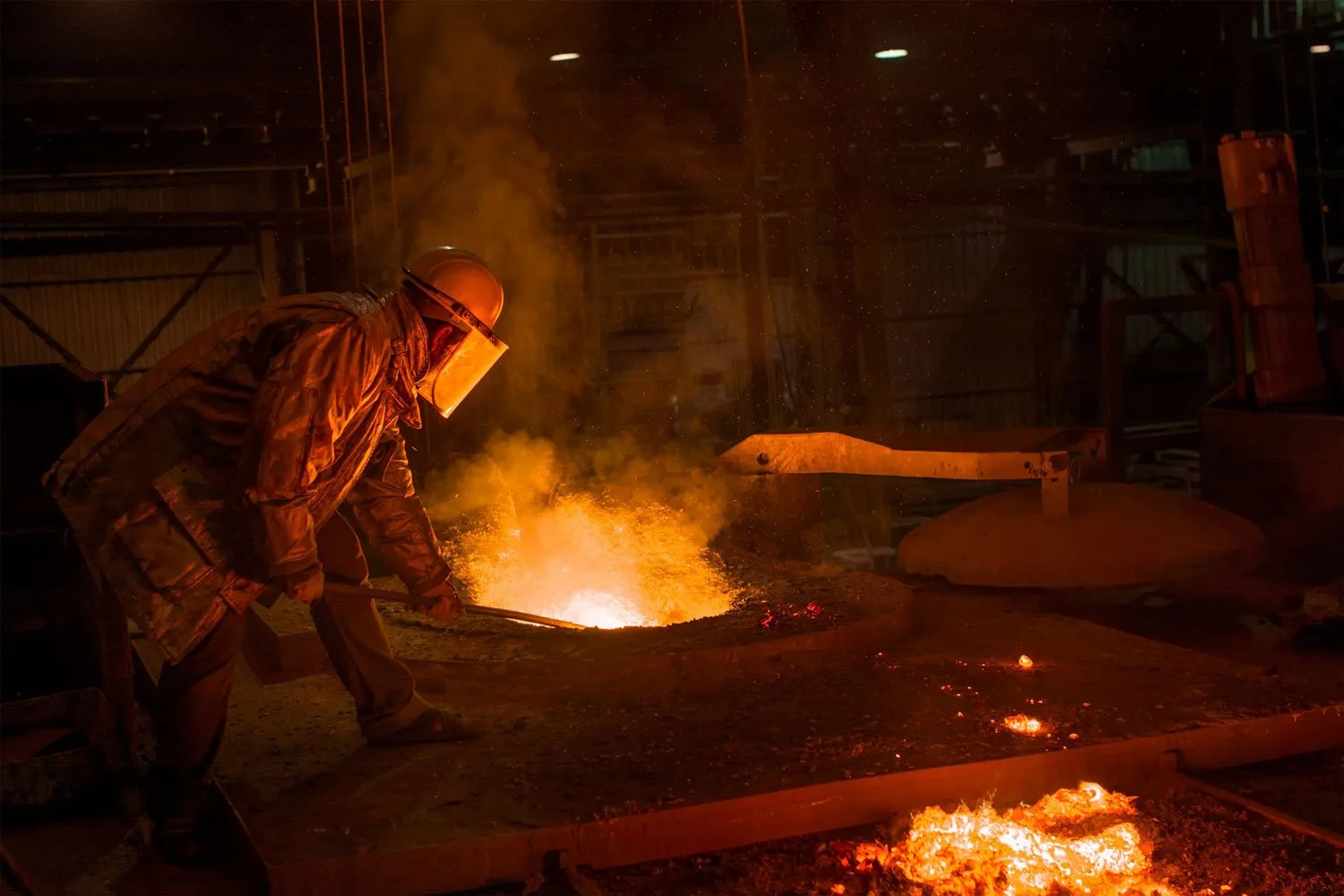 A worker in protective gear using a tool to manage molten metal in a foundry.