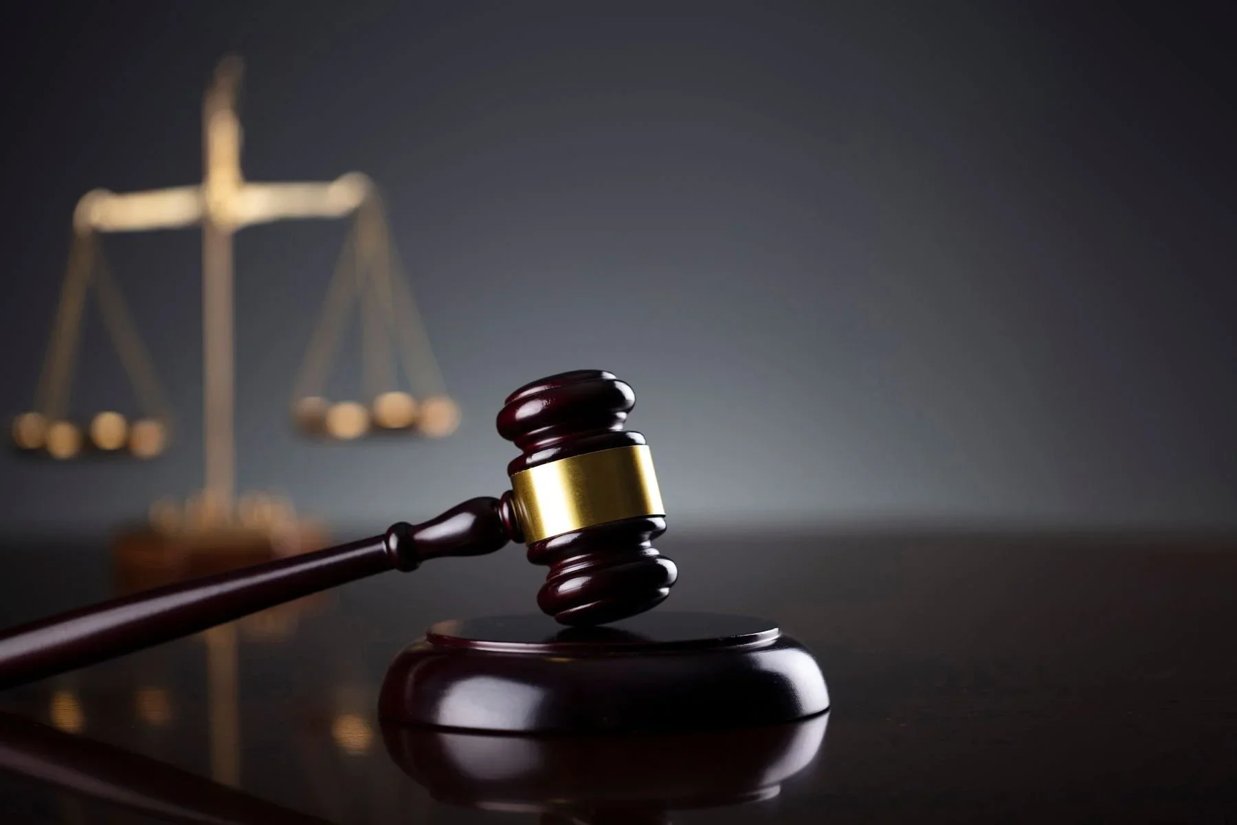 A wooden gavel with a gold band resting on a sound block, with a blurred set of scales of justice in the background.