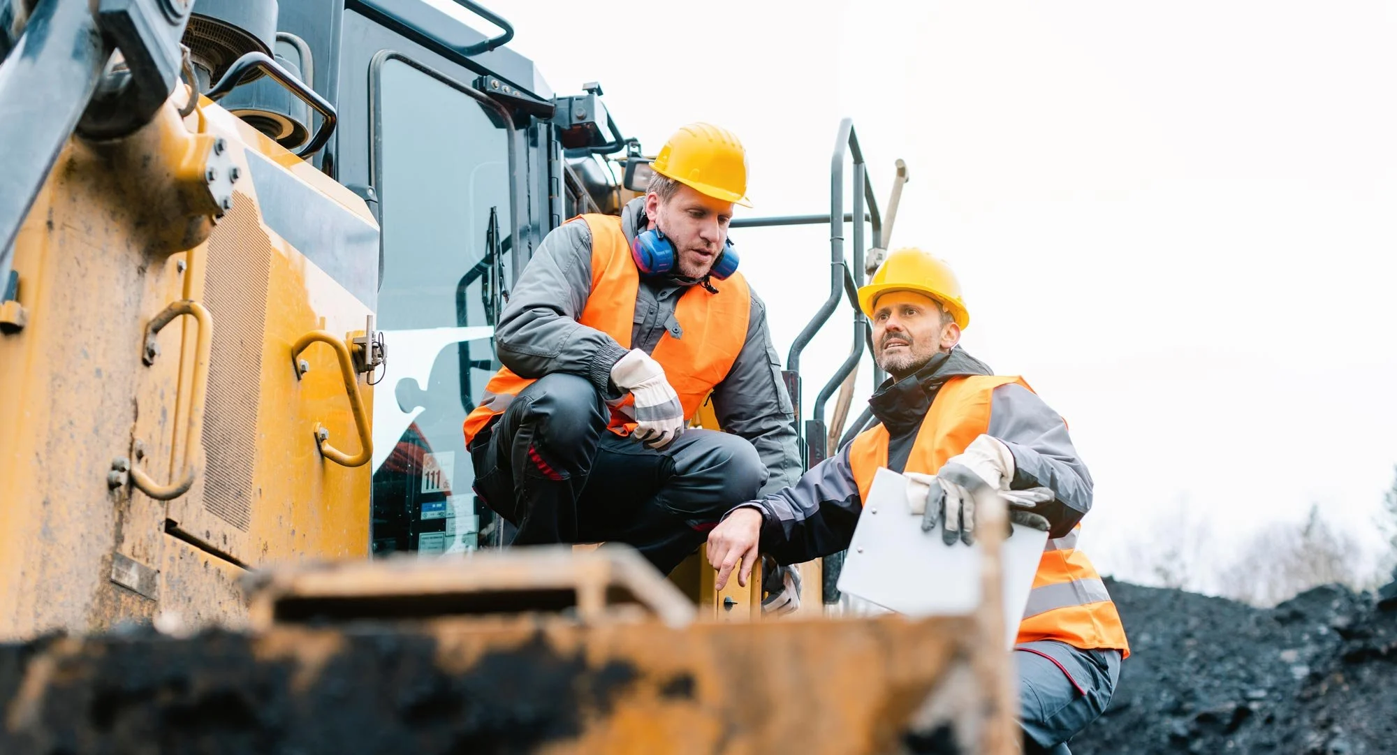 Two consultants in orange vests and yellow hard hats discuss next to a large construction vehicle.