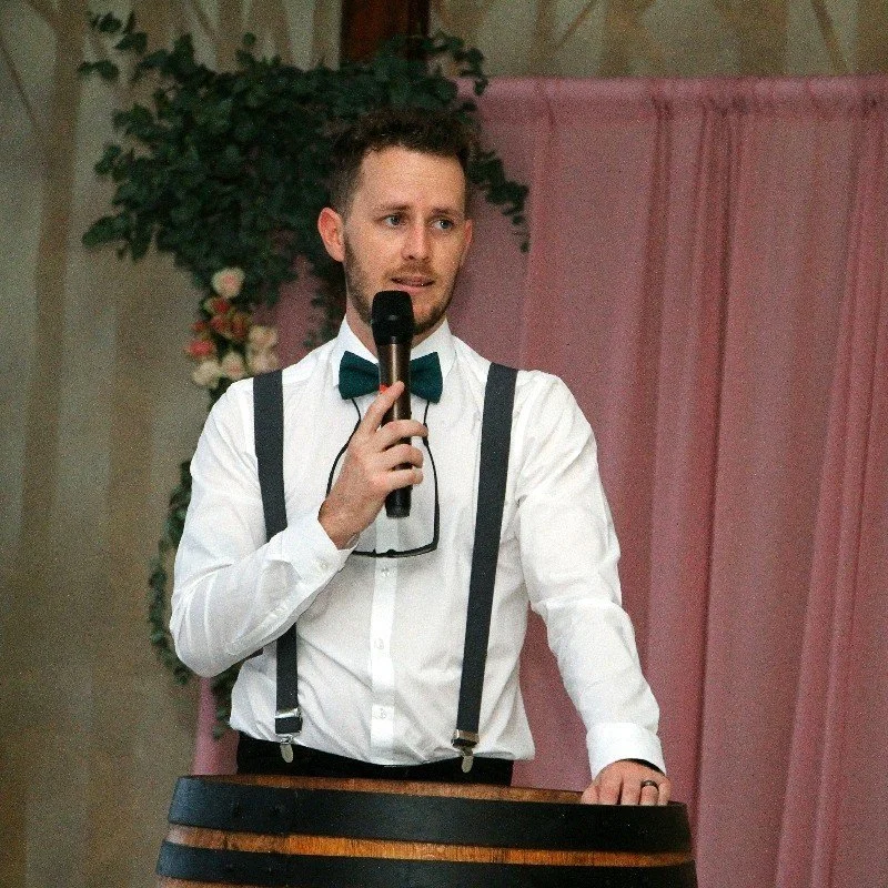 A man in a white shirt, teal bow tie, and suspenders speaking into a microphone at a formal event with a pink curtain and floral arrangement in the background.