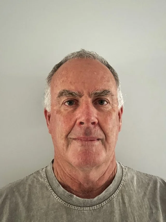 A middle-aged man with short gray hair and wearing a gray shirt, standing against a plain light-colored wall.