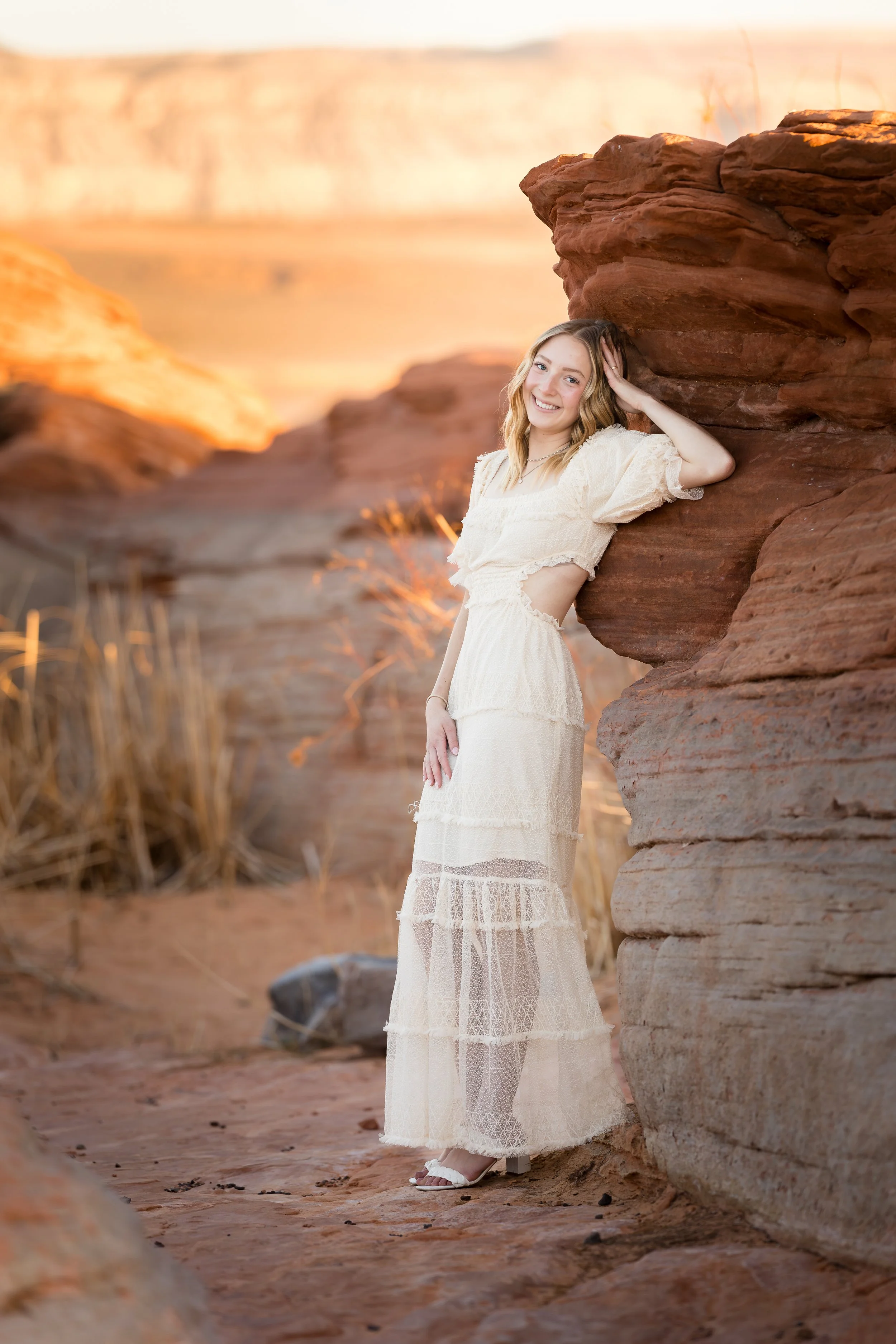 A senior girl wearing a white dress, posing against red rocks with her elbow bent and with sand hollow lake in the back ground along with golden plants and red sand, facing the camera