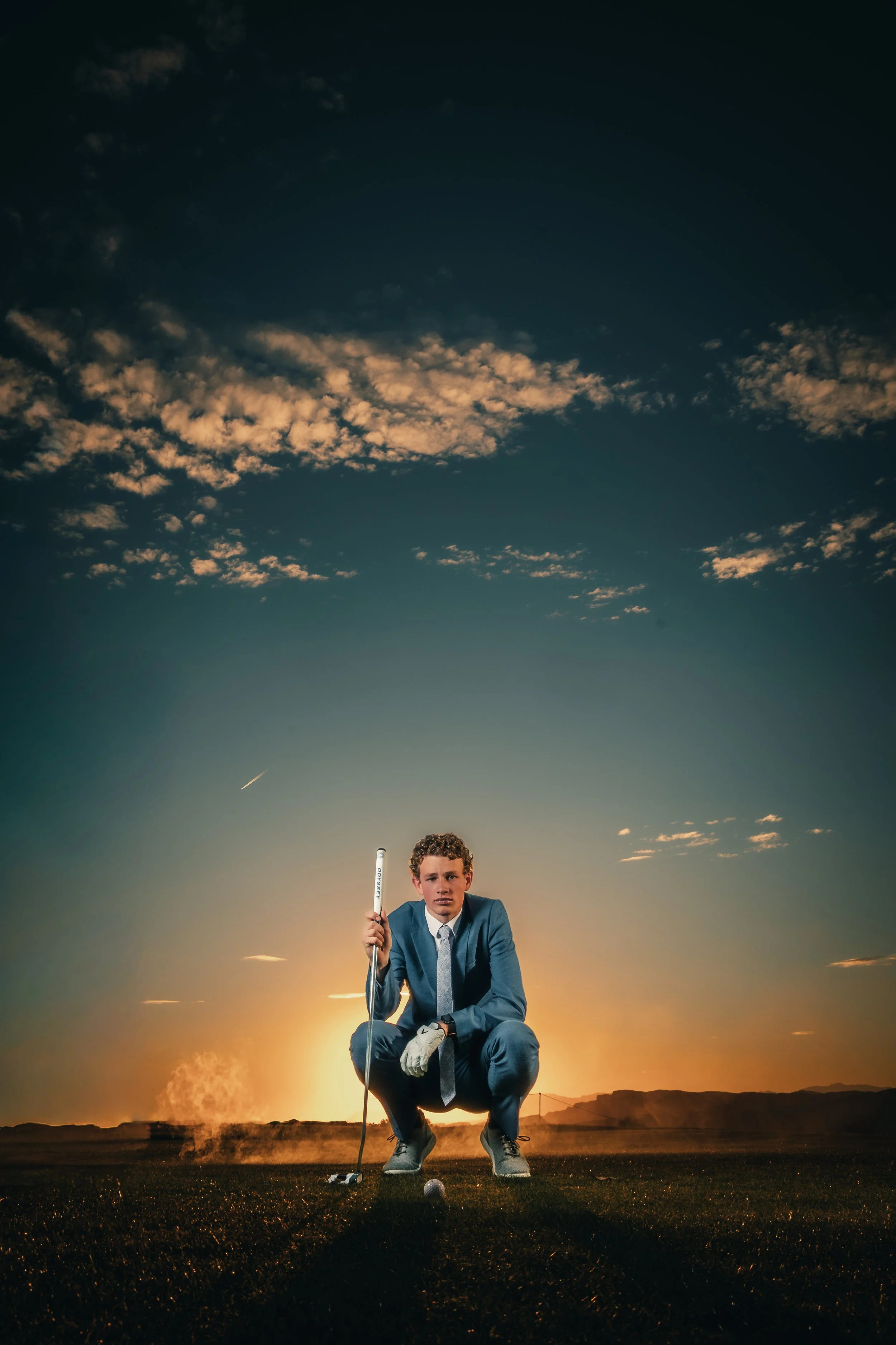 A man dressed in a suit and tie crouches on a golf course at sunset, holding a golf club, with the sky and clouds illuminated by the setting sun in the background.
