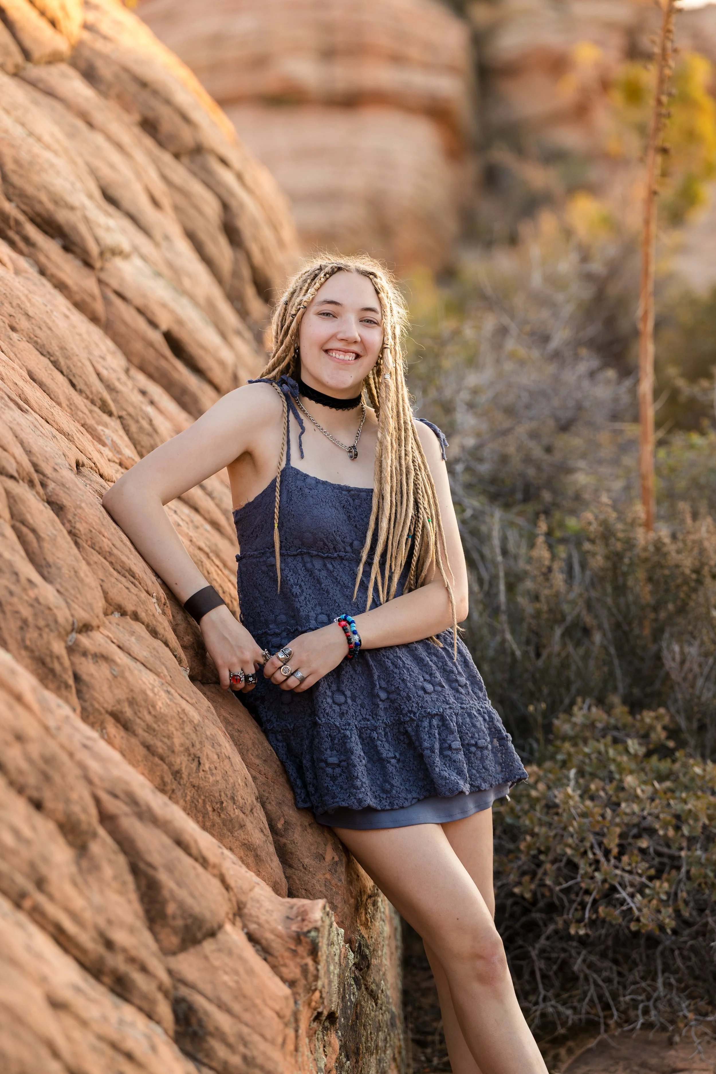 a blond senior girl with long braids, wearing a blue summer dress and neck choker, leaning against rippled red rocks with desert plants in the background