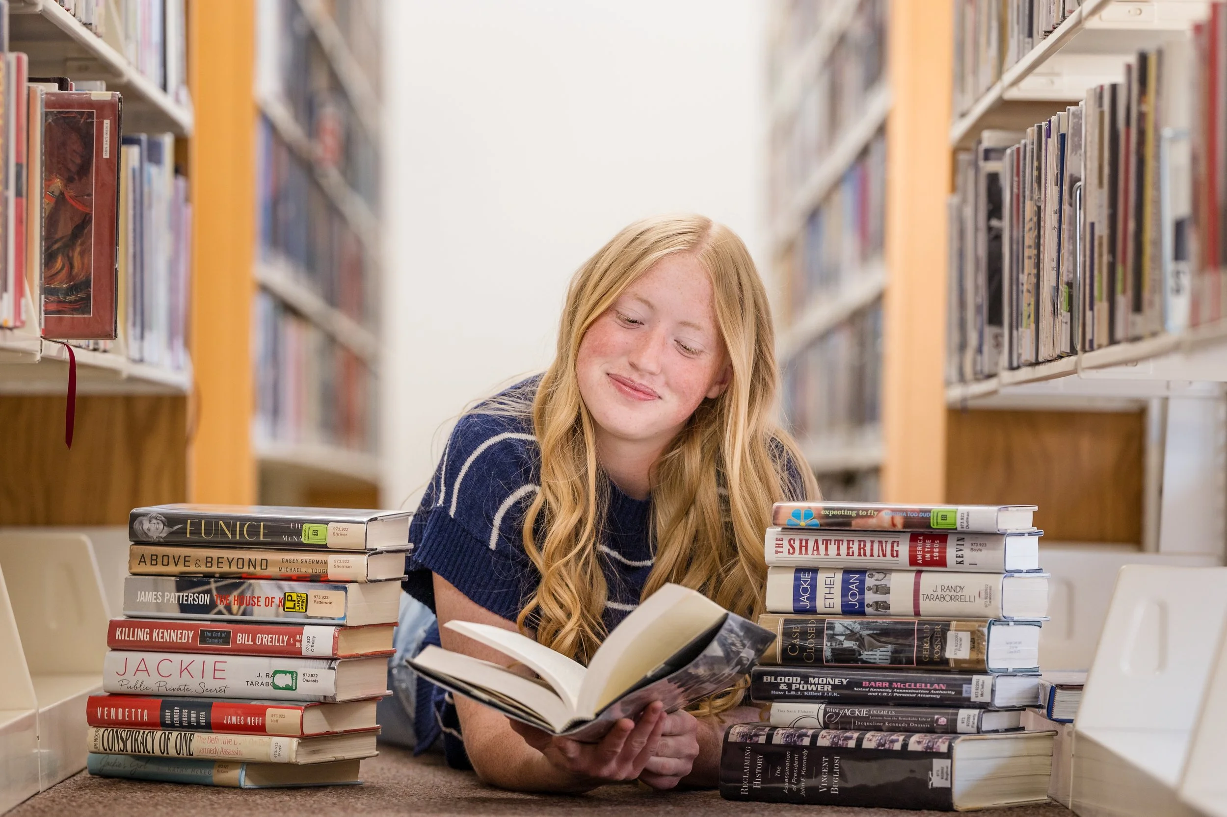A high school senior girl with blond long hair, wearing a striped blue shirt, laying on the floor of a library surounded with books while looking at a book she appears to be reading