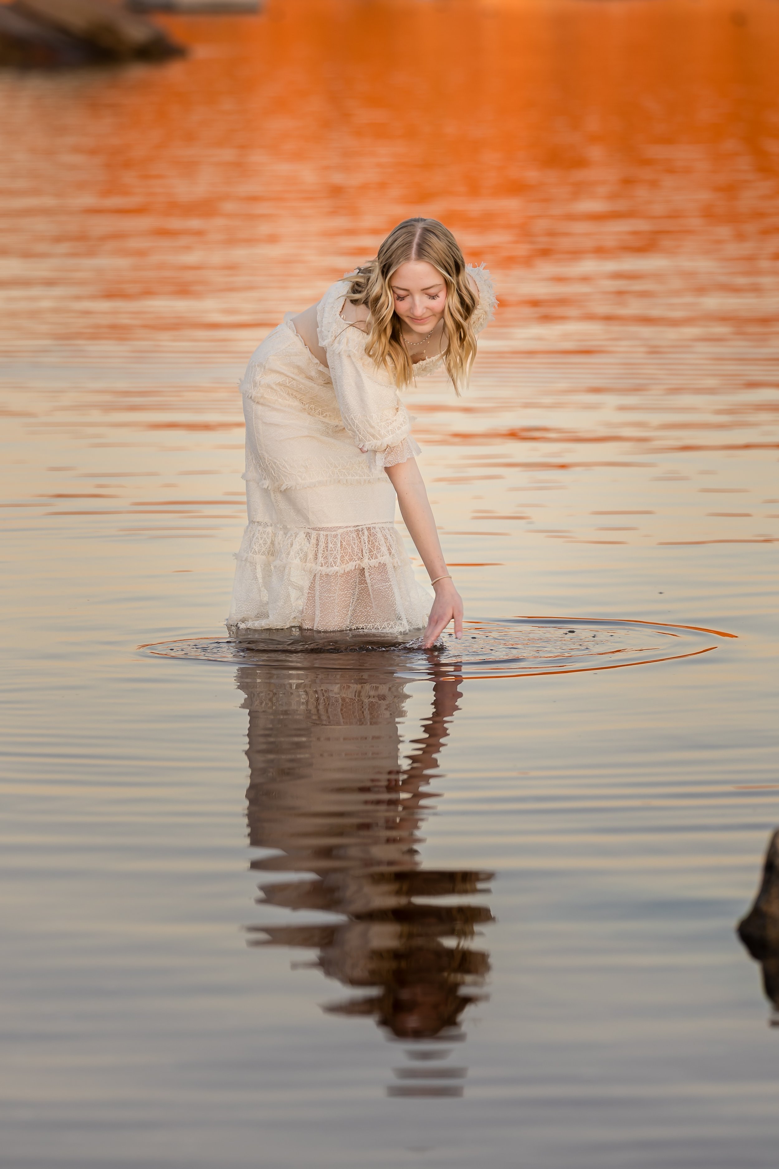 A woman in a white dress wading in a calm body of water during sunset with orange and pink sky reflections.