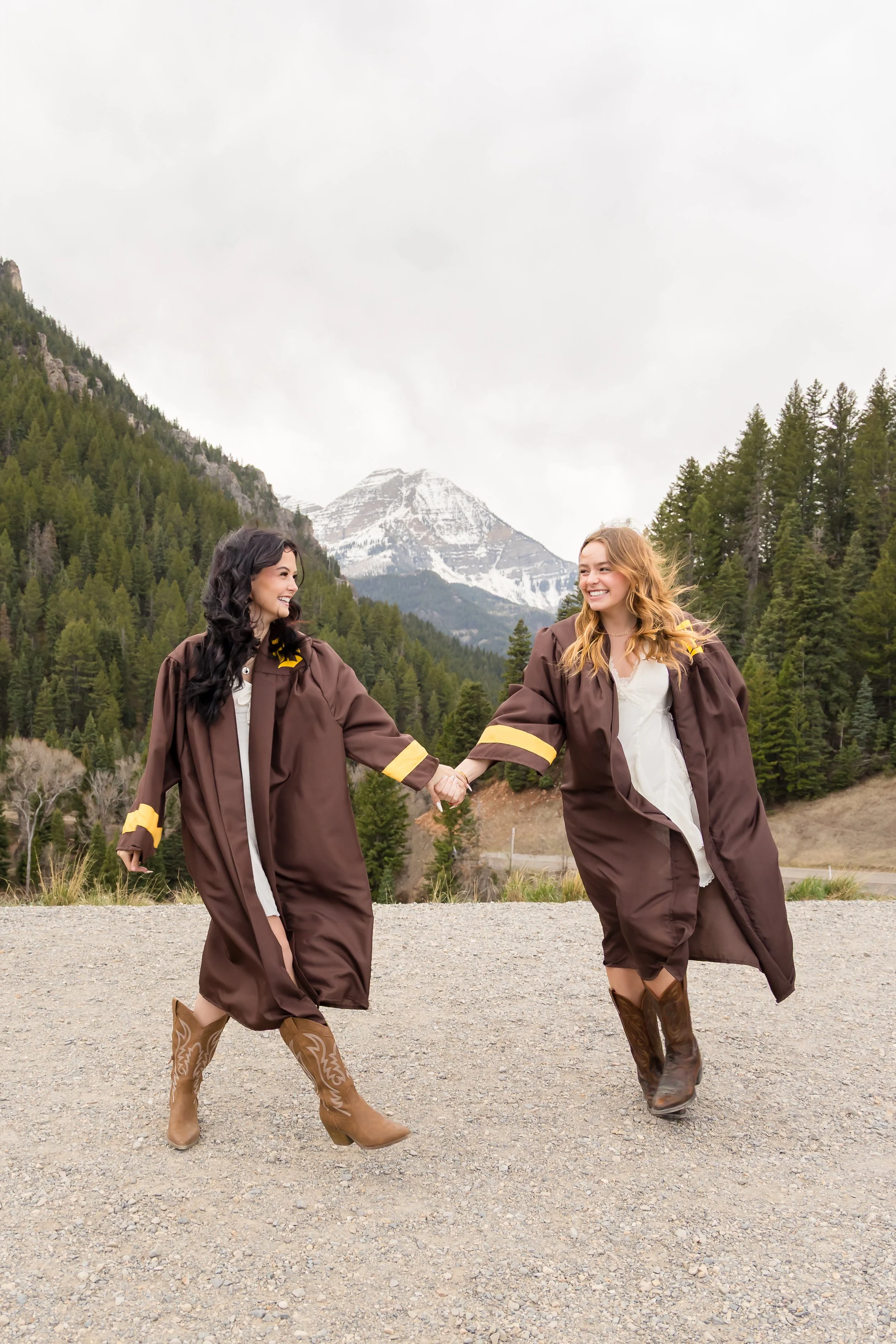 Two women in graduation gowns holding hands and smiling outdoors with mountains, trees, and overcast sky in the background.