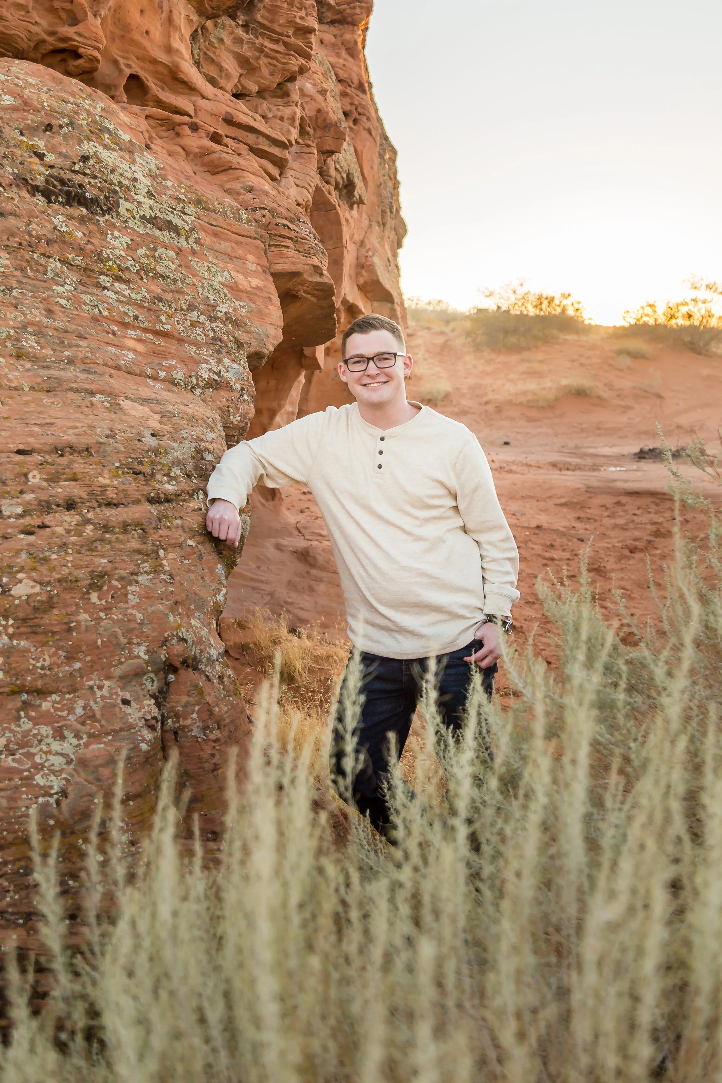 A young man wearing glasses, a beige long-sleeve shirt, and dark pants standing outdoors against a red rock formation during sunset. He is smiling and leaning against the rock with one arm.