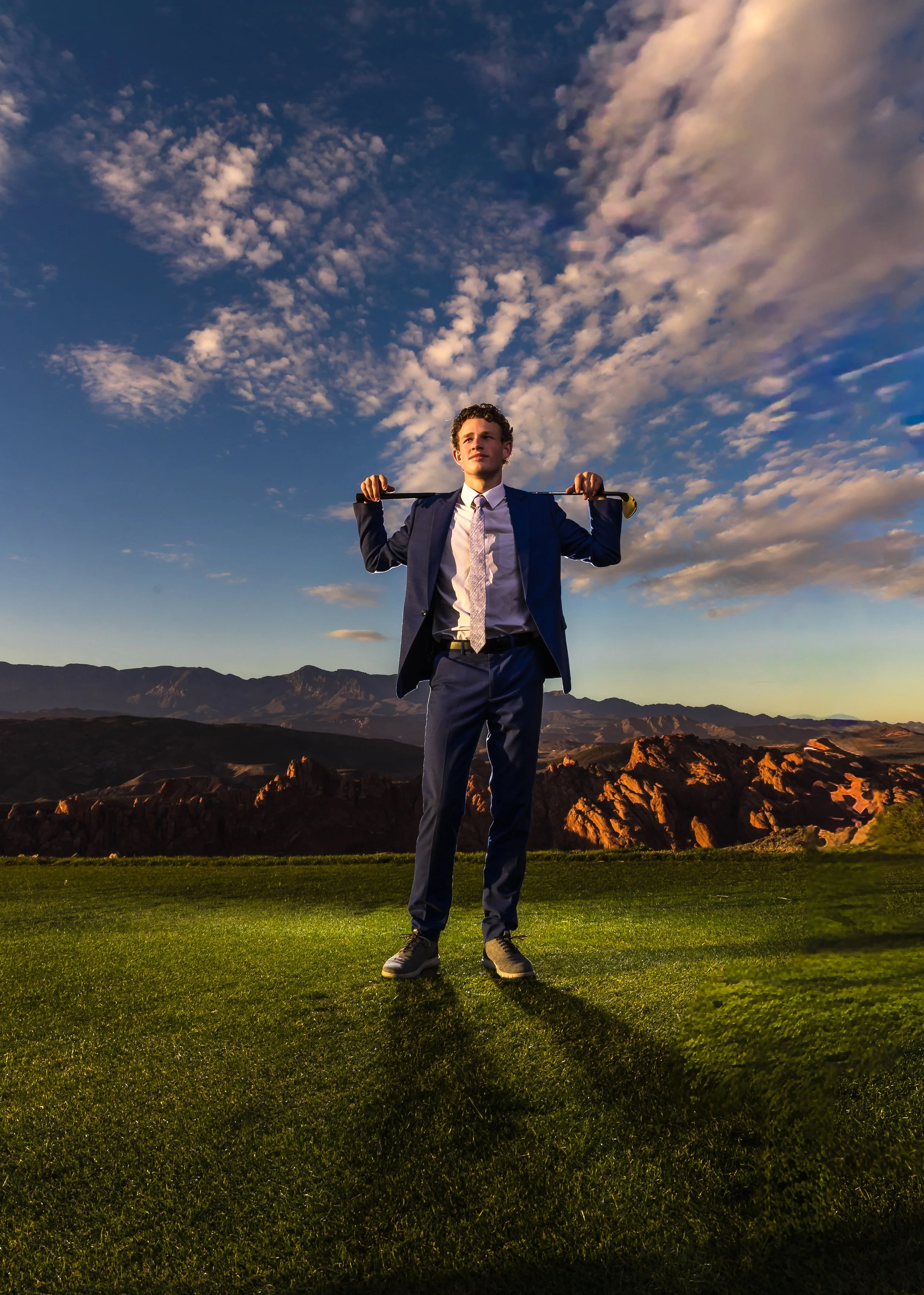 A young man in a suit holding a broomstick over his shoulders, standing on a grassy field at sunset with mountains and a partly cloudy sky in the background.