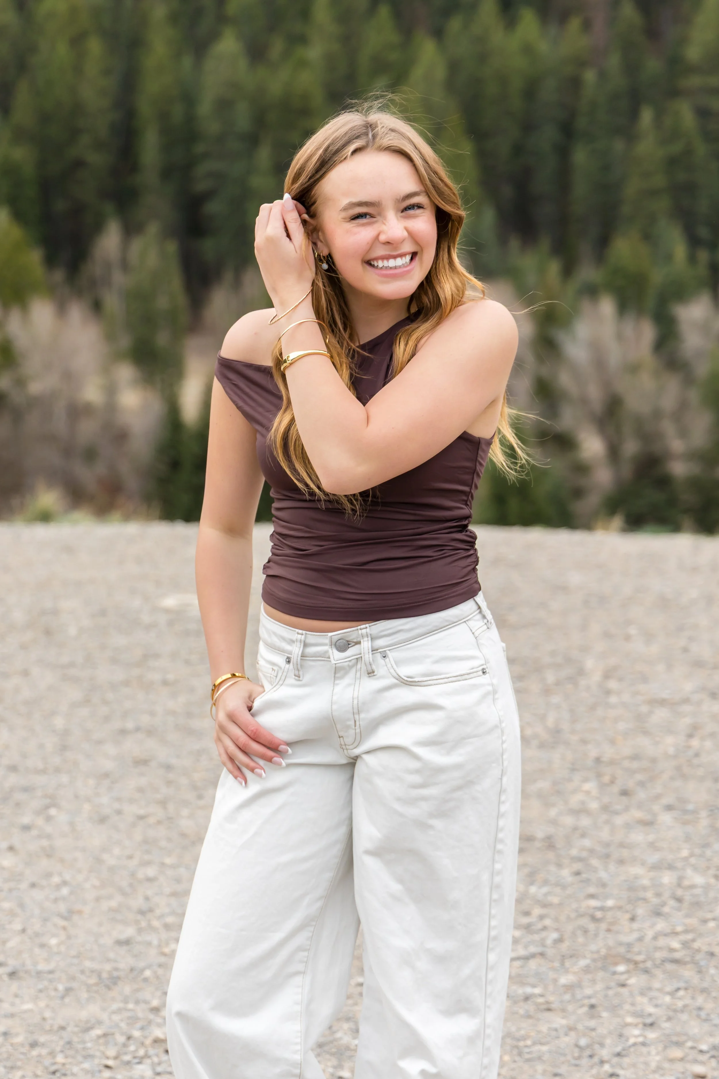 a light haired senior girl wearing a brown shirt and jeans, tucking her hair behind her ear while she laughs at the camera with evergreen trees behind her