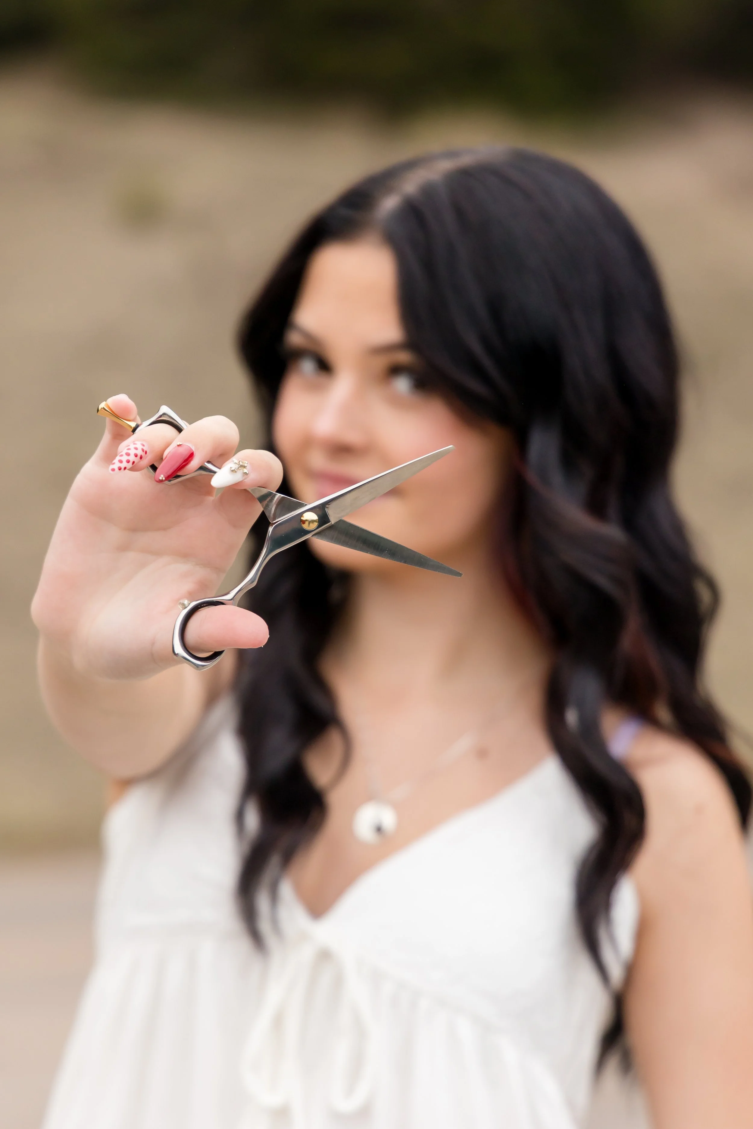 A woman with black wavy hair holds a large pair of scissors close to the camera, with a neutral outdoor background.
