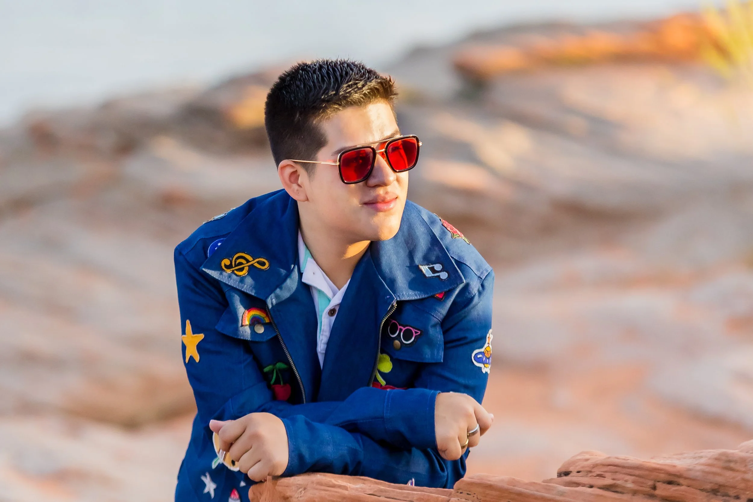 a senior boy wearing a blue denim jacket with lots of patches and red sunglasses leaning on a red rock with the lake in the back ground