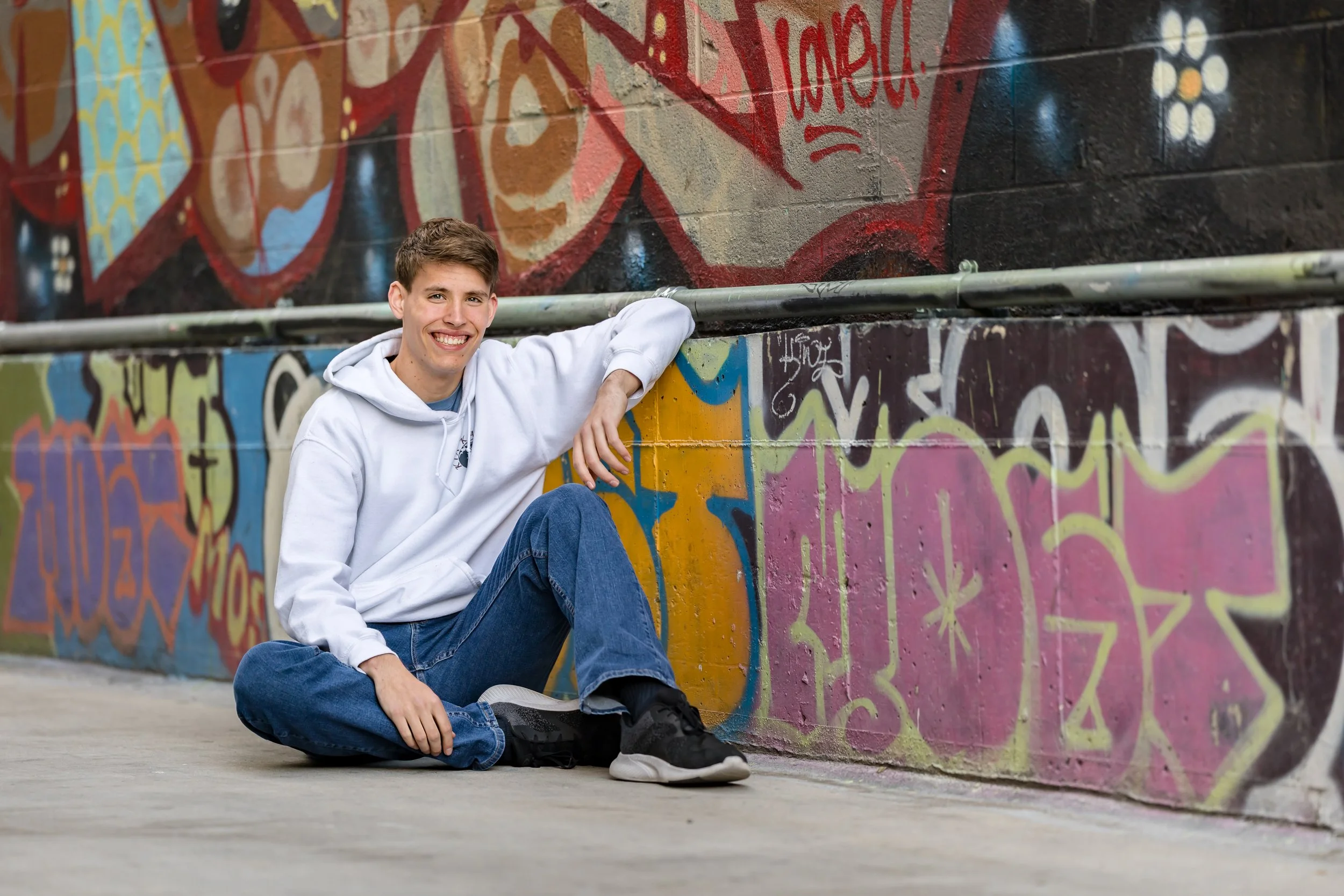 A young man sitting on the ground beside a graffiti-covered wall, smiling and looking at the camera. He is wearing a white hoodie and blue jeans.