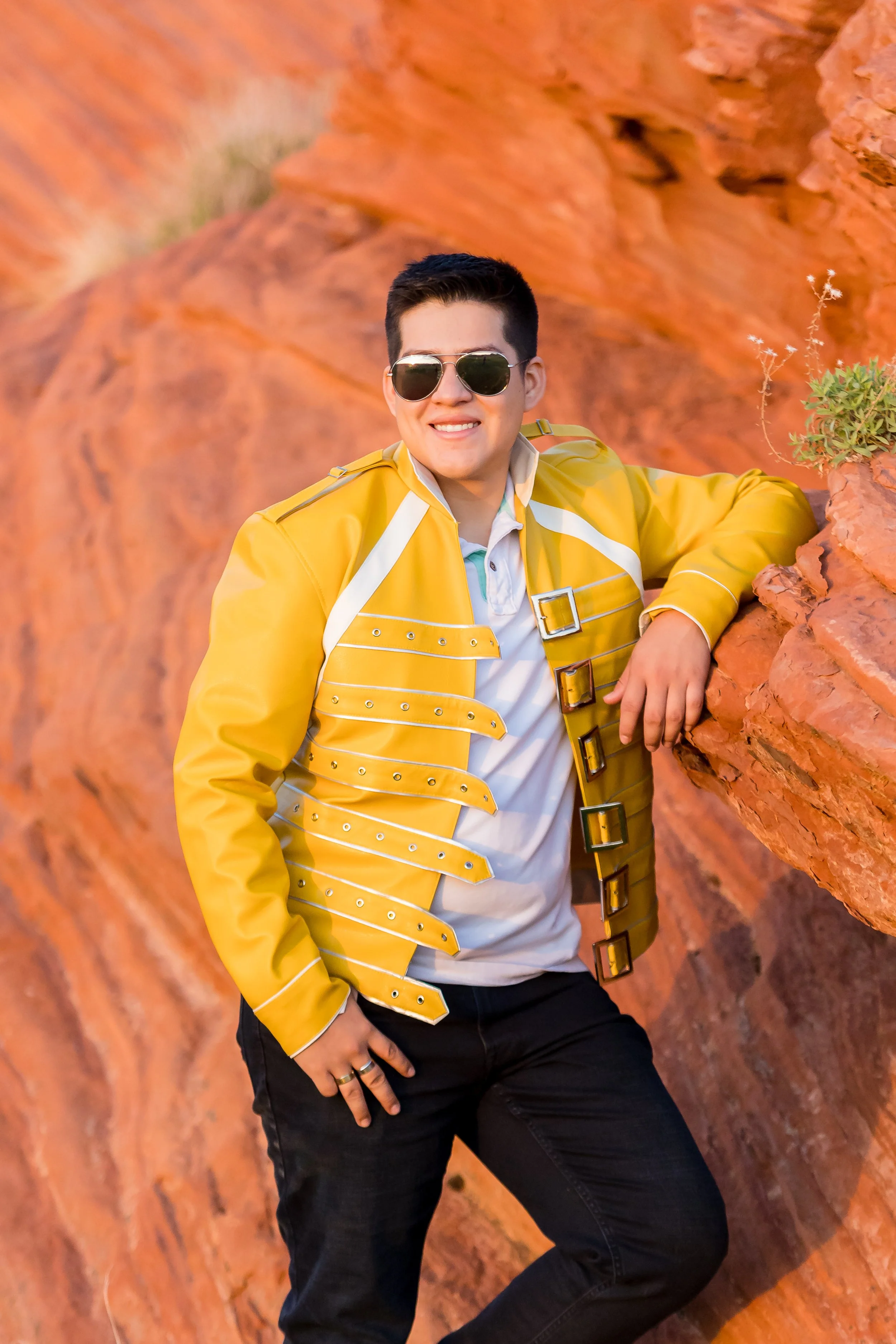 A young man with short dark hair wearing sunglasses, a white t-shirt, and a yellow jacket with buckle details, posing in front of red rock formations with a small green plant nearby.