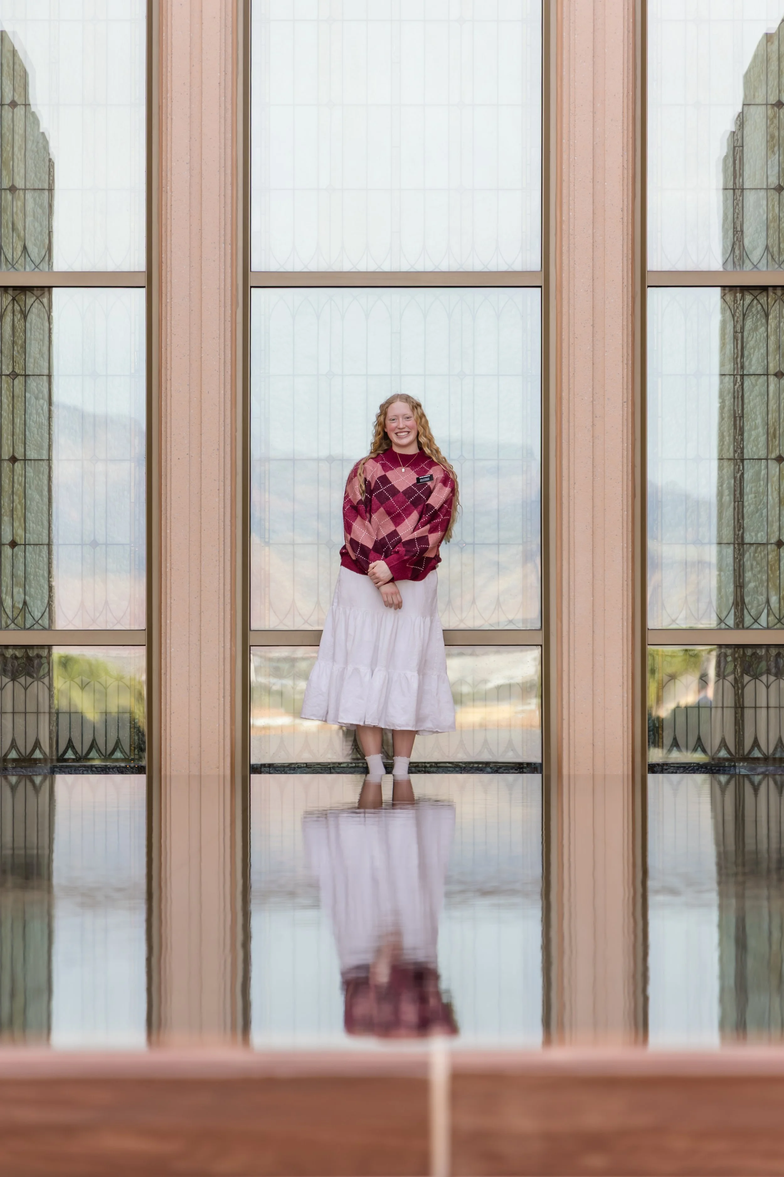 A young woman standing outside an LDS Temple with large stained-glass windows, smiling, wearing a red and pink argyle sweater, a white skirt, and white socks.
