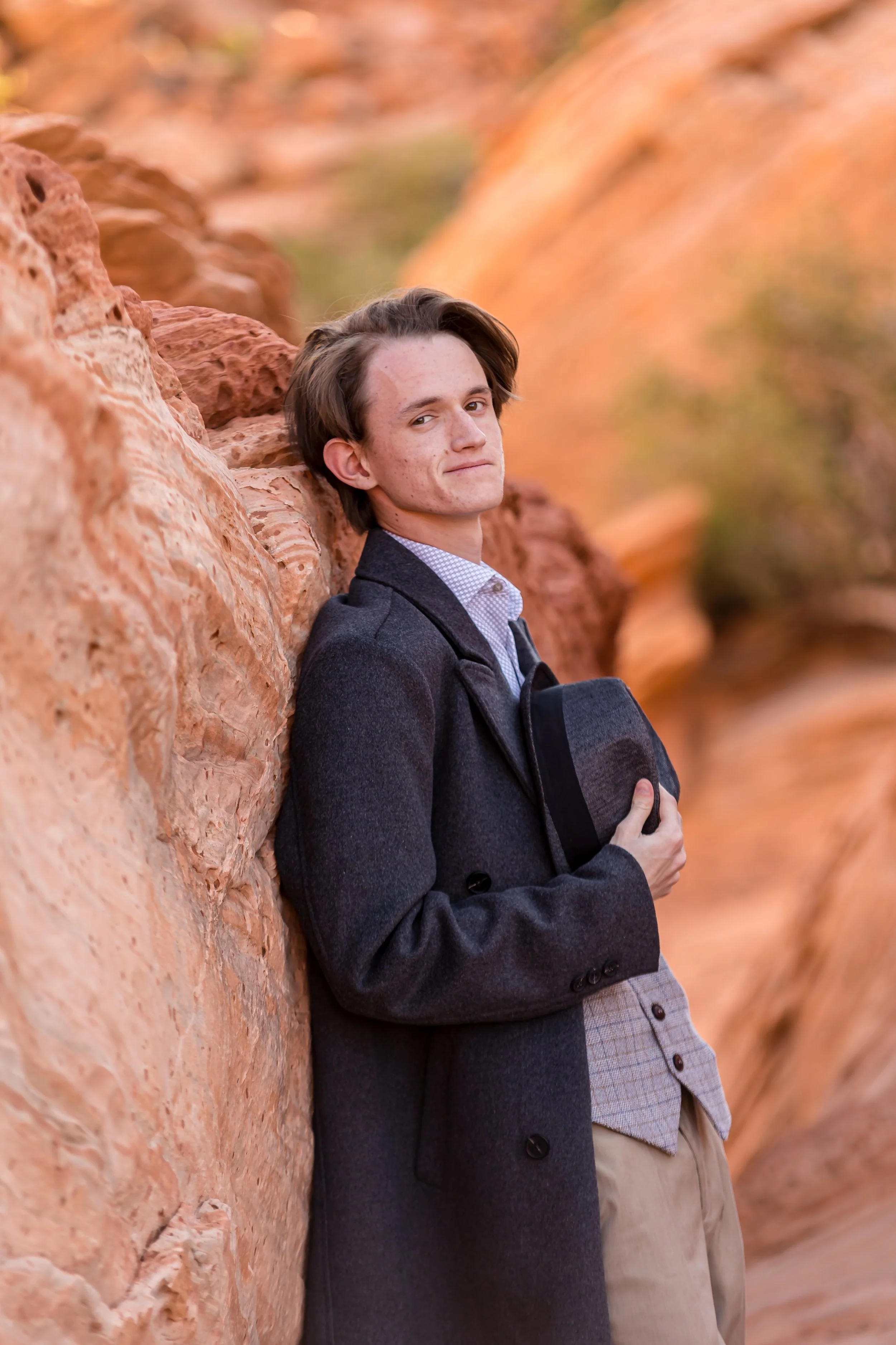 dark haired Senior guy wearing a coak and vest leaning agianst red rock and holding a hat to his chest while smiling at the camera