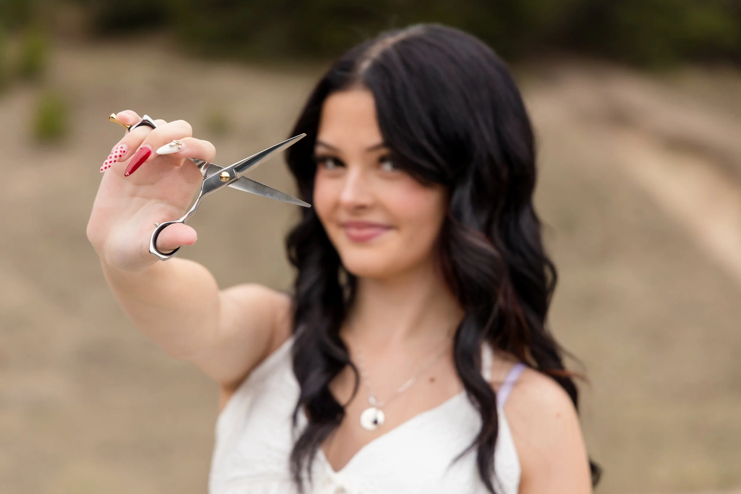 a dark haired senior girl wearing a white tank top, looking at the camera, slightly out of focus, while holding a pair of hair cutting shears that are in focus
