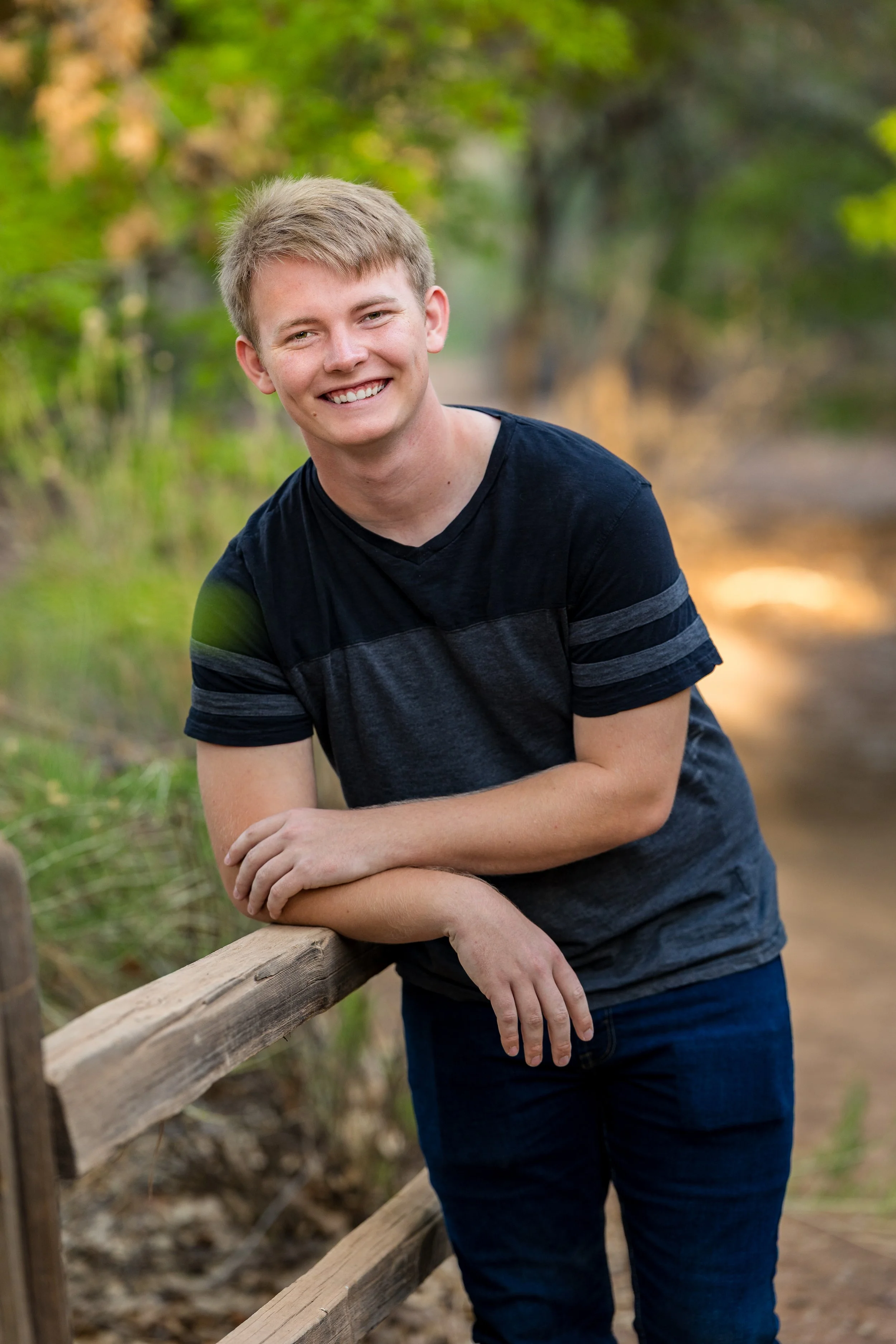 Blonde senior guy leaning agianst a wood fence wearing dark colors with green trees and sunlight behind him