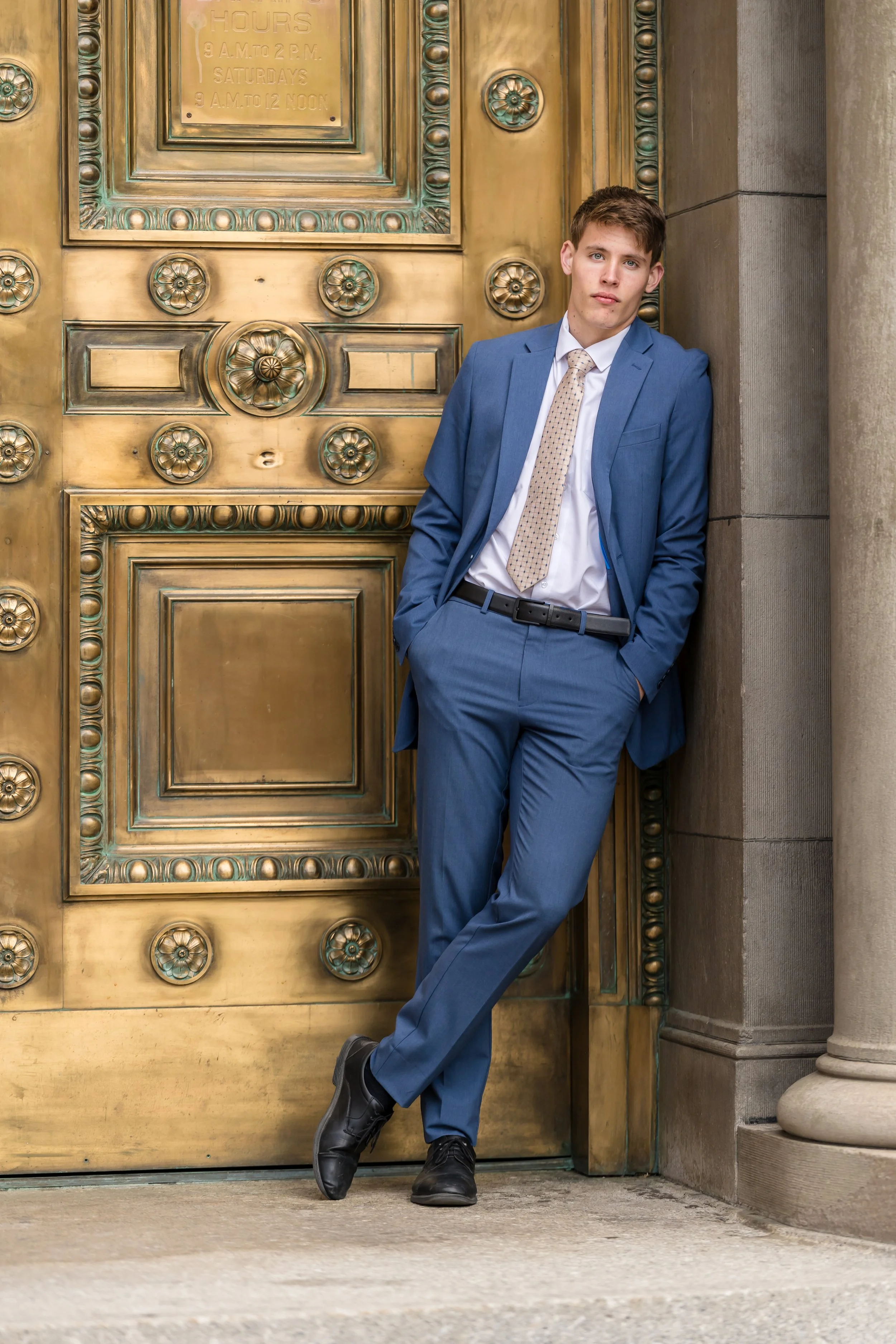 A young man in a blue suit leaning against a large, ornate gold door with his hand in his pocket and looking at the camera.