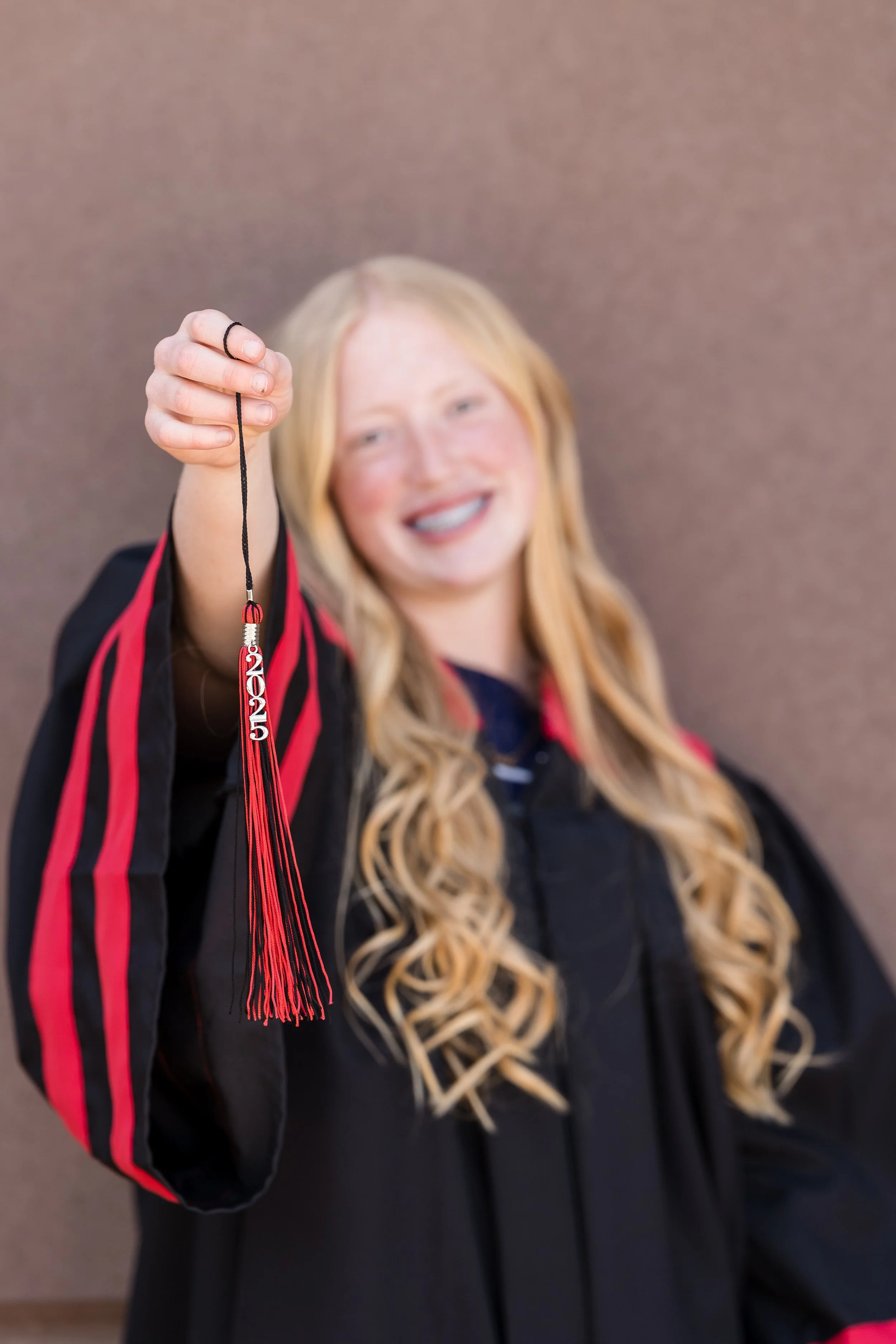 A smiling woman with long blonde hair in a graduation gown, holding a red and black tassel.