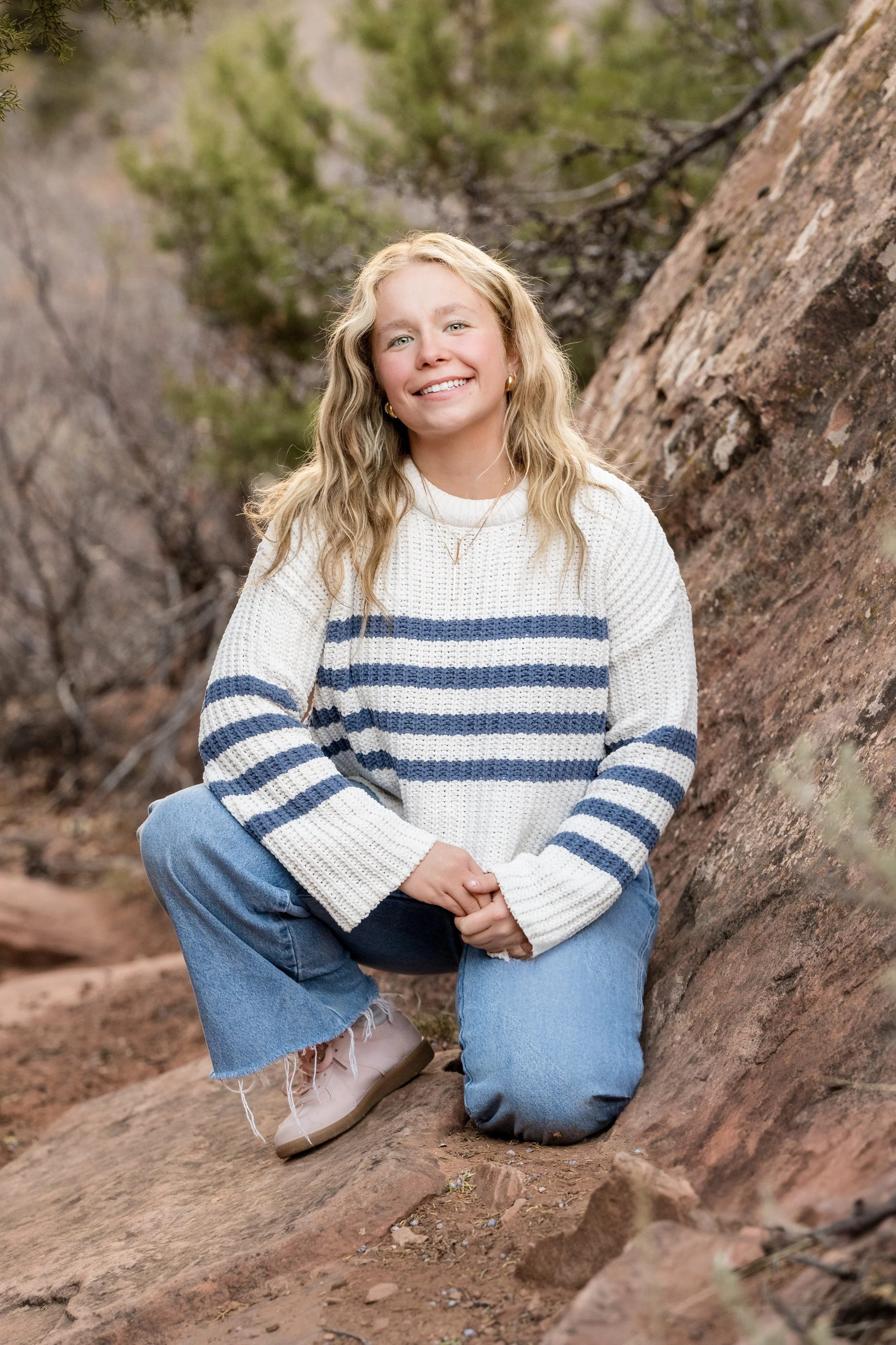 Blonde female senior wearing a striped white sweater crouching near some rocky terain