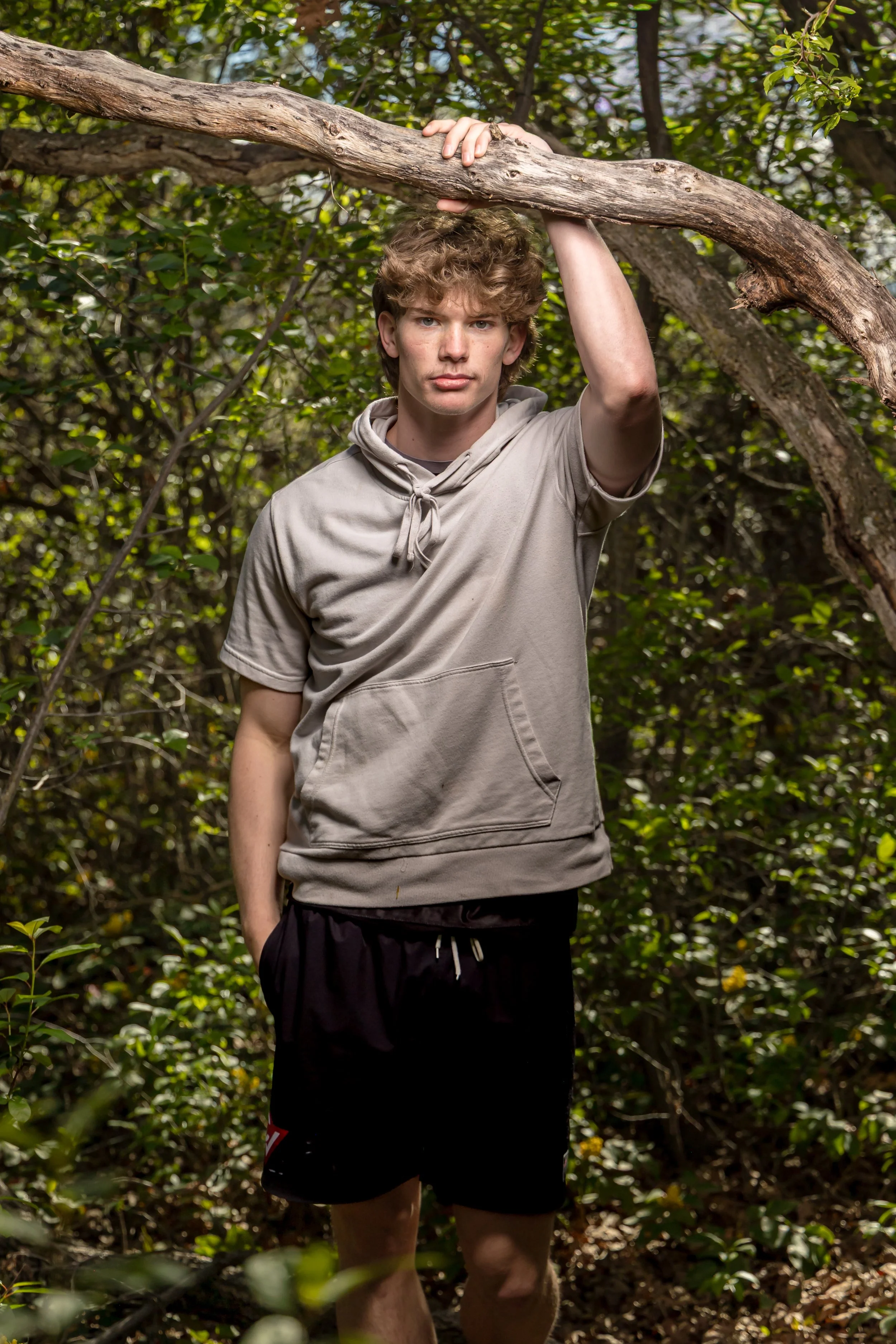 A young man with curly brown hair standing in a wooded area during daytime, holding onto a large tree branch with one hand, wearing a light-colored hoodie and black shorts.