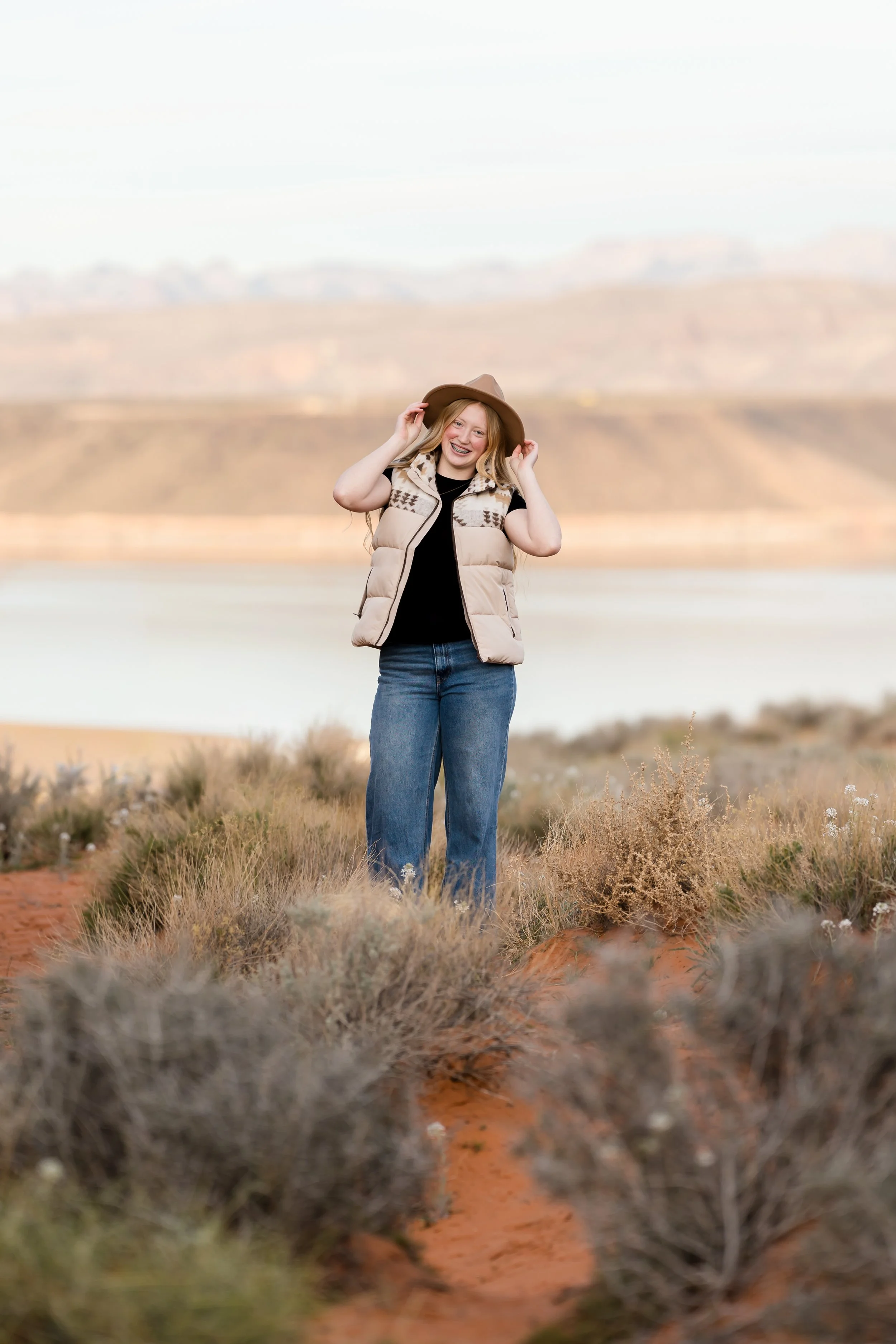 A young woman wearing a beige vest, black shirt, blue jeans, and a brown hat standing on a desert landscape with shrubs and mountains in the background, smiling and holding her hat with both hands.