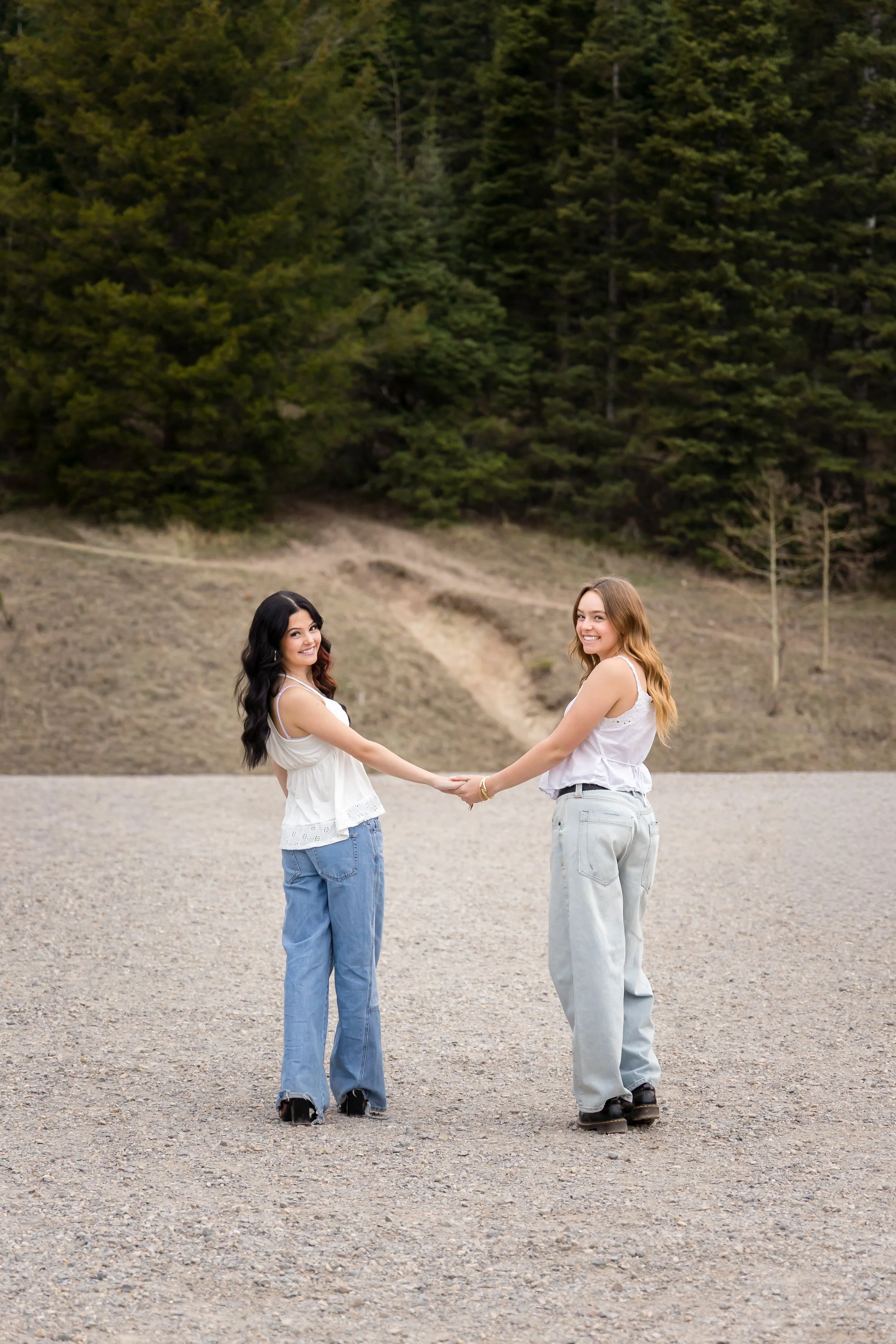 Two young women holding hands and smiling outdoors with a forested hillside in the background.