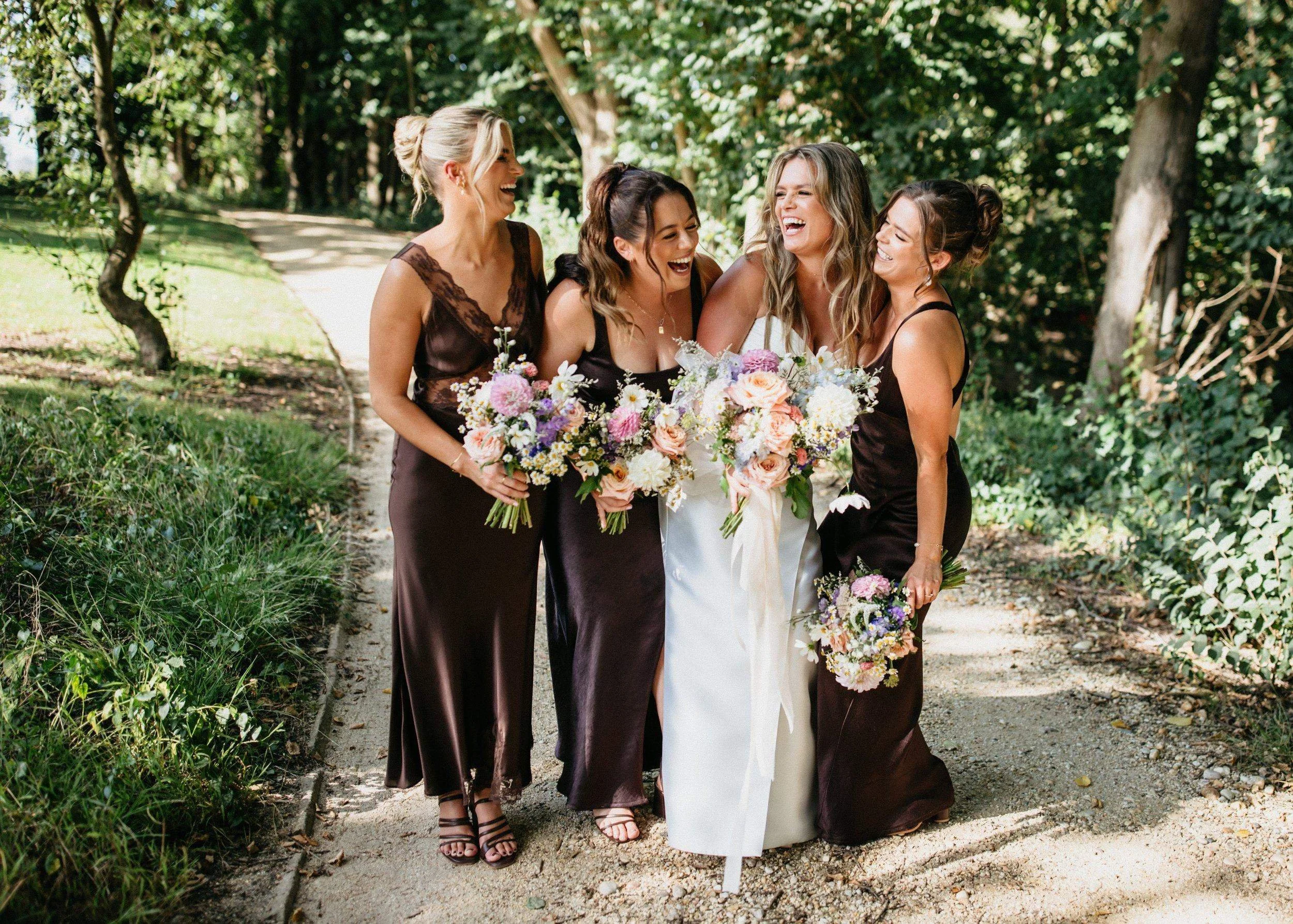 Group of five women at a wedding, standing on a dirt path in a wooded area, all smiling and laughing, with one woman in a white dress holding a bouquet of flowers.