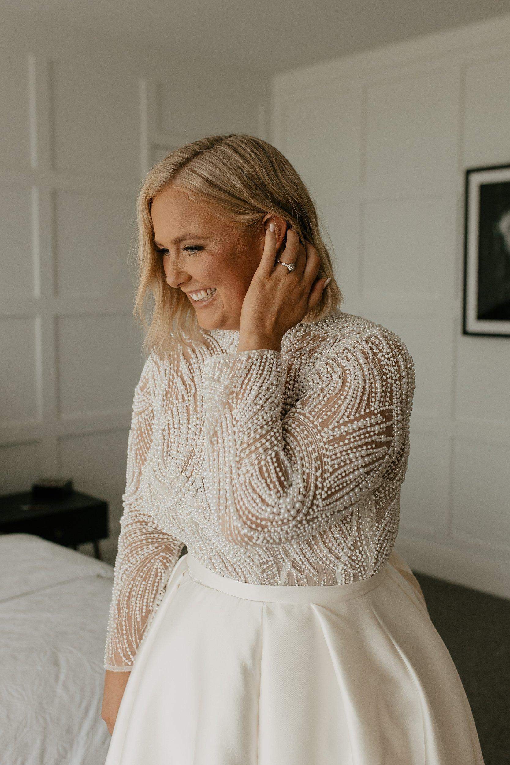 A woman smiling and touching her ear, dressed in a beaded, long-sleeve top with a full, white satin skirt.