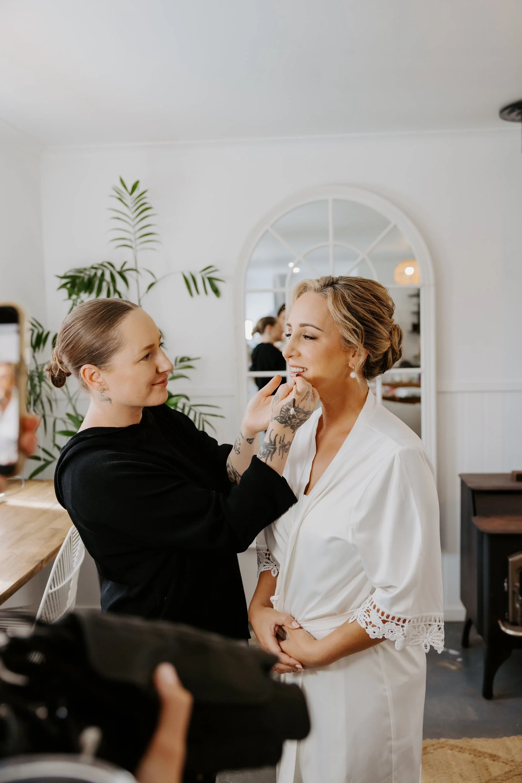 A woman in a white dress is being makeup touched up by a makeup artist in a studio or salon with a large mirror in the background and green plants.