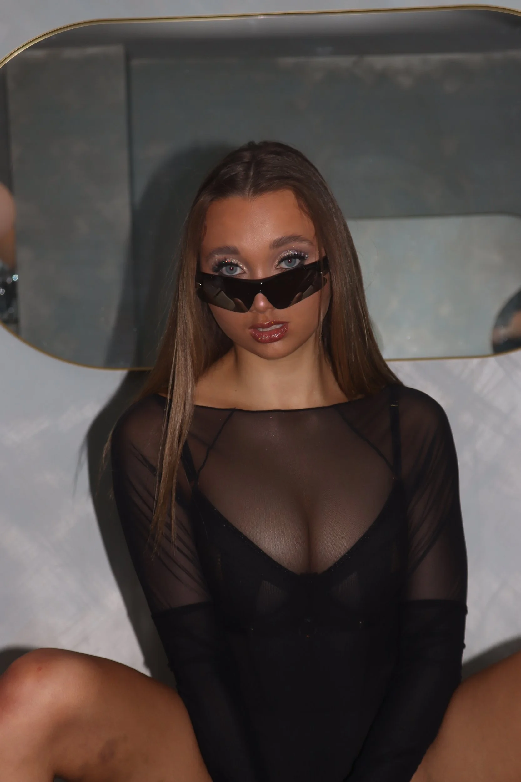 A woman with long brown hair wearing black sunglasses and a sheer black outfit, sitting in front of a mirror.