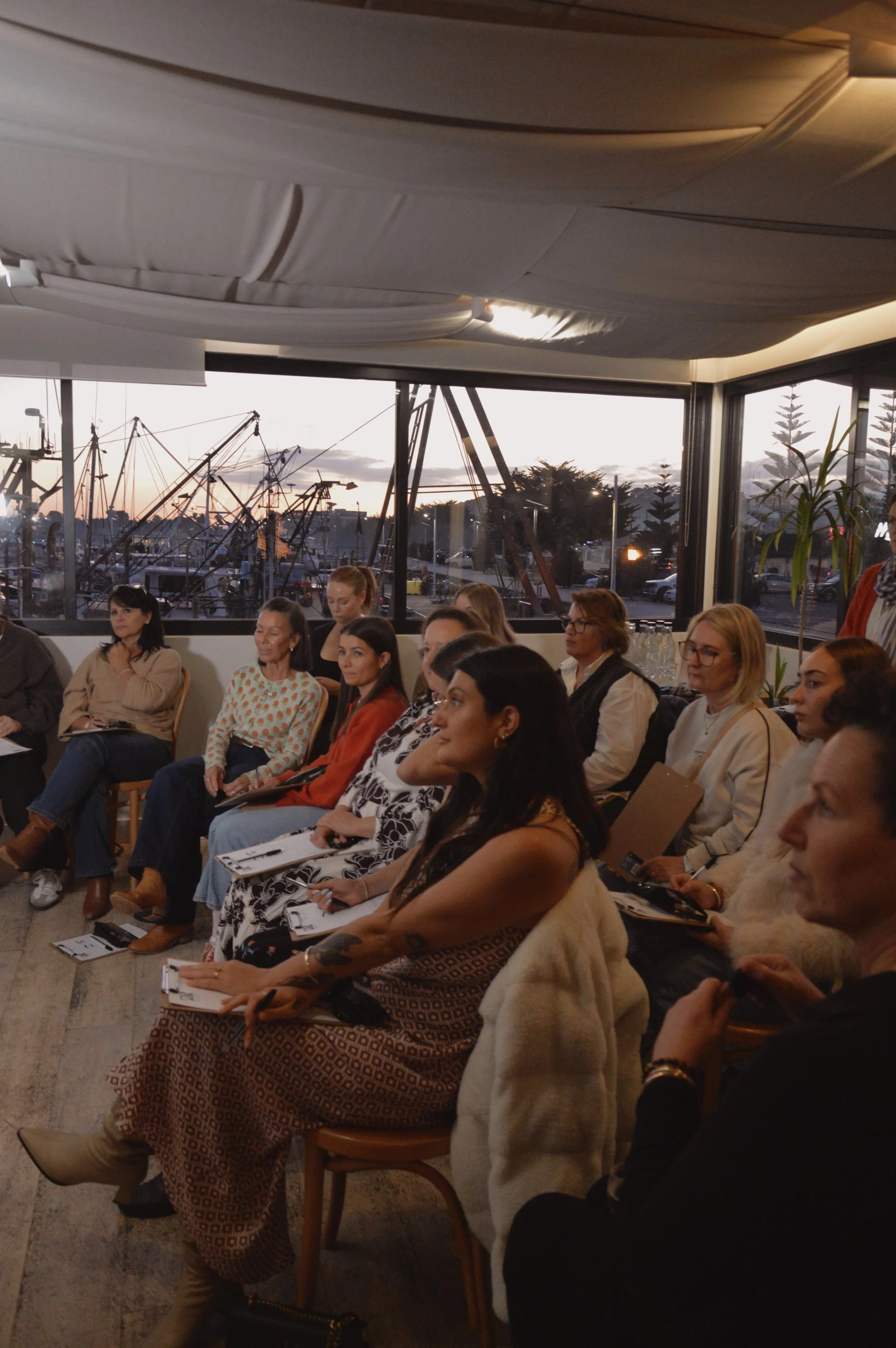A group of people sitting attentively at a meeting or conference in a room with large windows revealing a marina with boats and a sunset sky in the background.