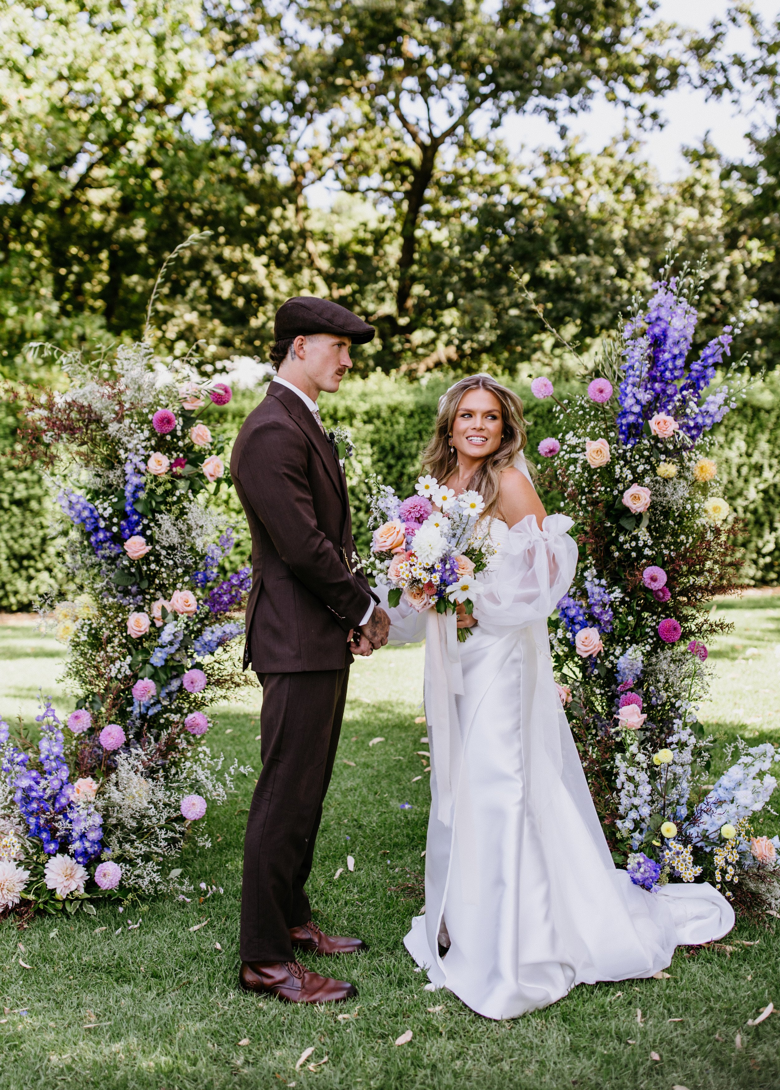 A bride and groom standing outdoors in front of a large floral arch at their wedding ceremony, holding hands and smiling.
