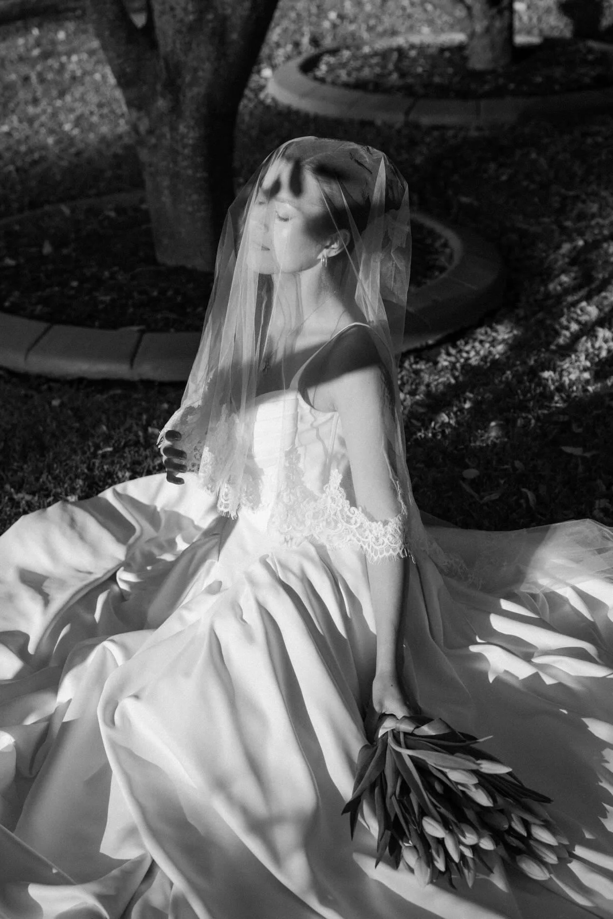 A woman in a wedding gown sitting outdoors in black and white, her face partially covered by a veil, with a bouquet of flowers lying on the ground nearby.