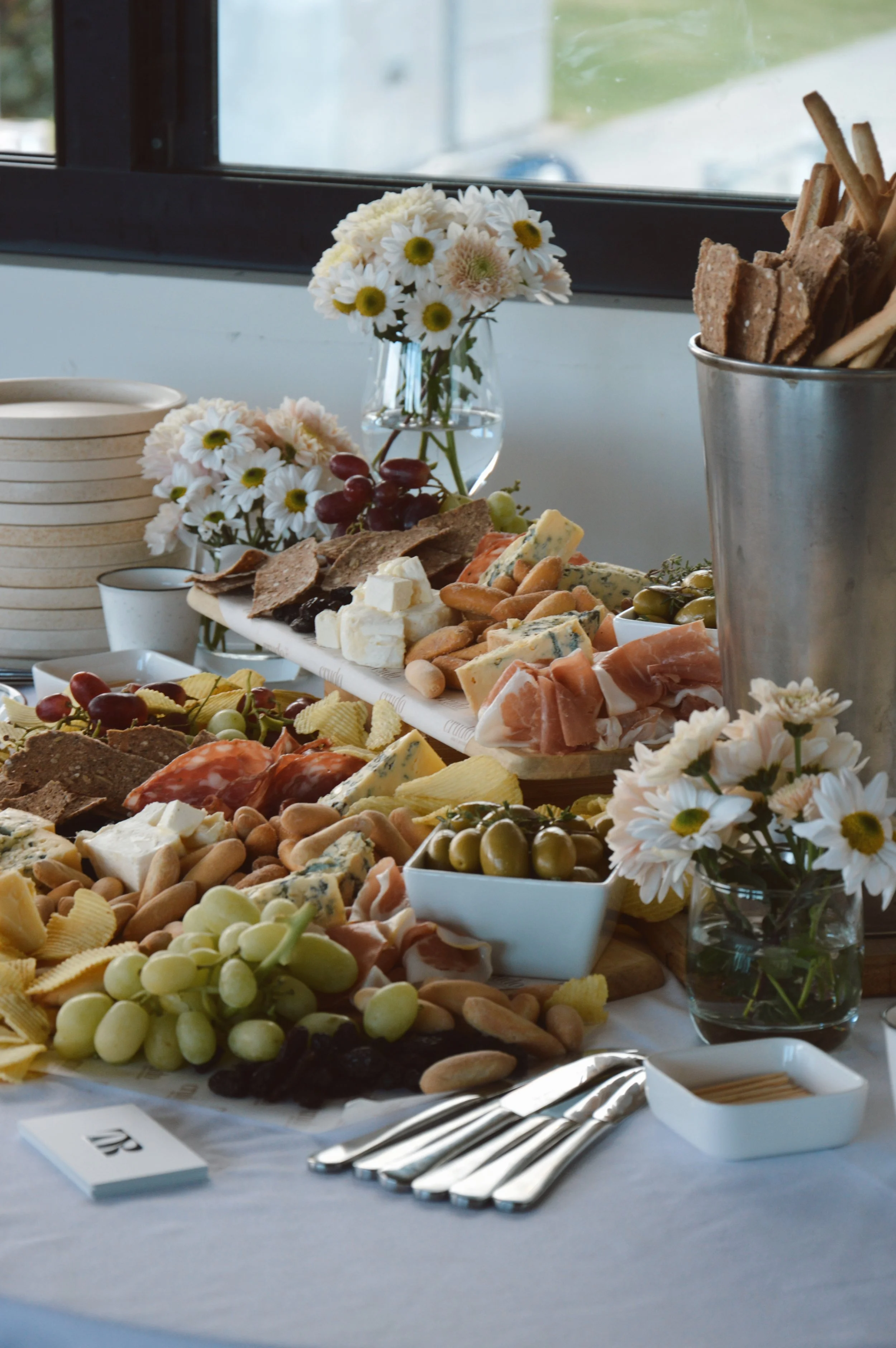A cheese and charcuterie platter with grapes, crackers, and various cheeses, decorated with white flowers in a glass vase, on a table near a window.
