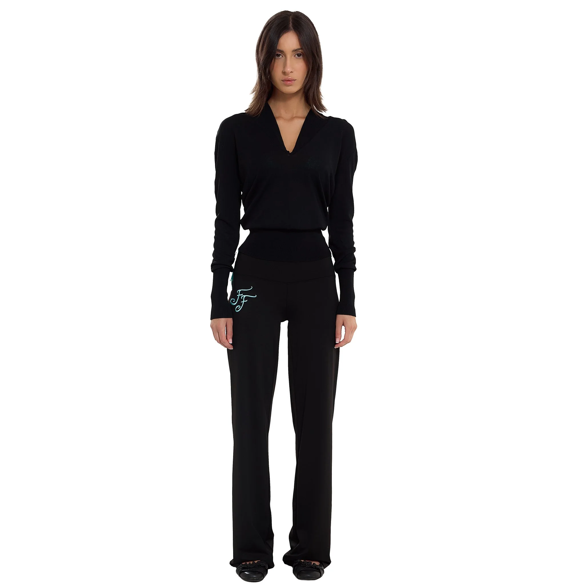 A woman standing against a plain white background, wearing black pants and a black long-sleeved top.