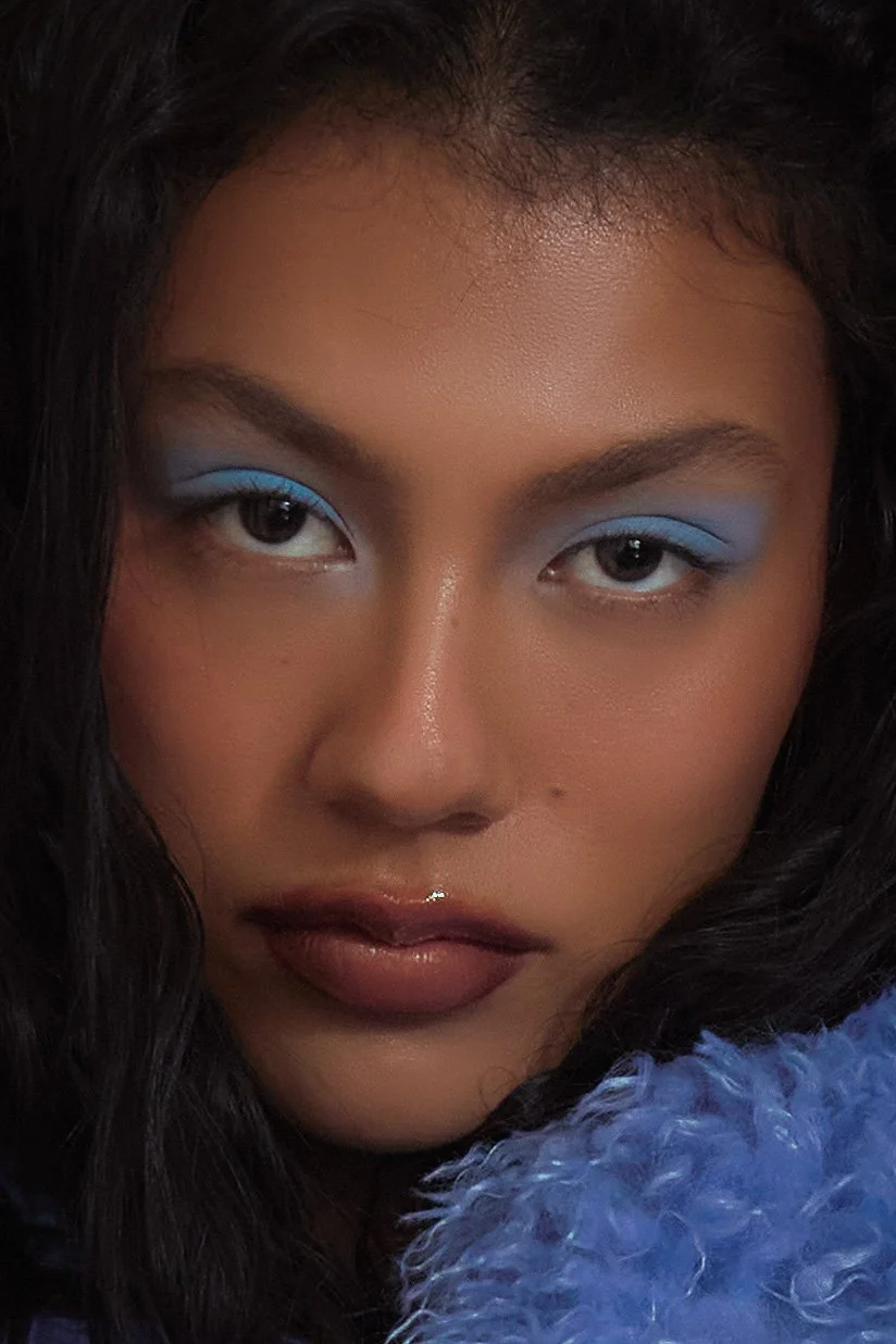 Close-up portrait of woman with medium skin tone, wavy dark hair, bold makeup with blue eyeshadow, and glossy lips, partially covered with a fluffy blue material.