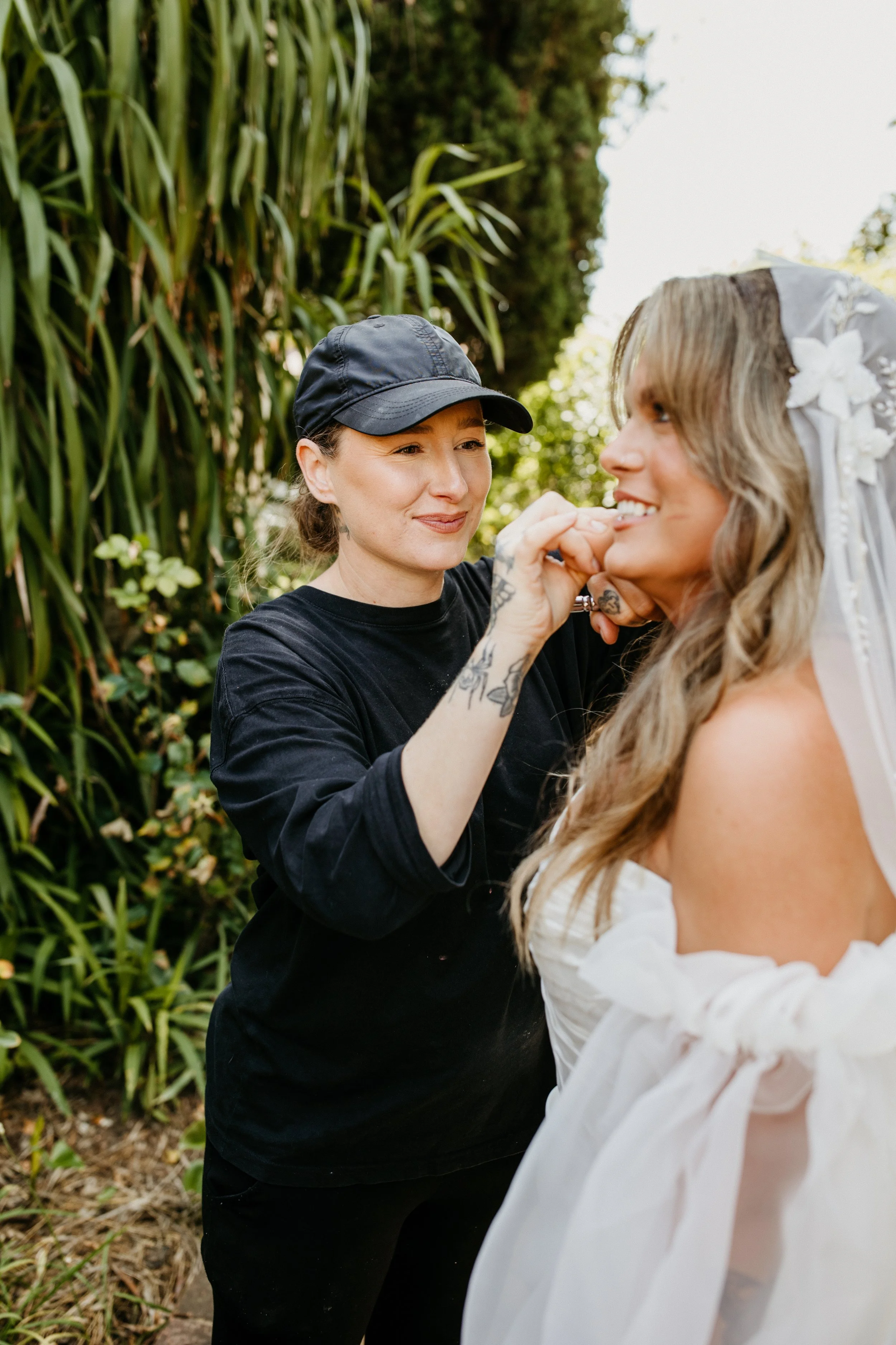 A makeup artist or stylist applies makeup or face paint to a smiling woman in a white dress, outdoors among greenery, with another woman in the foreground.